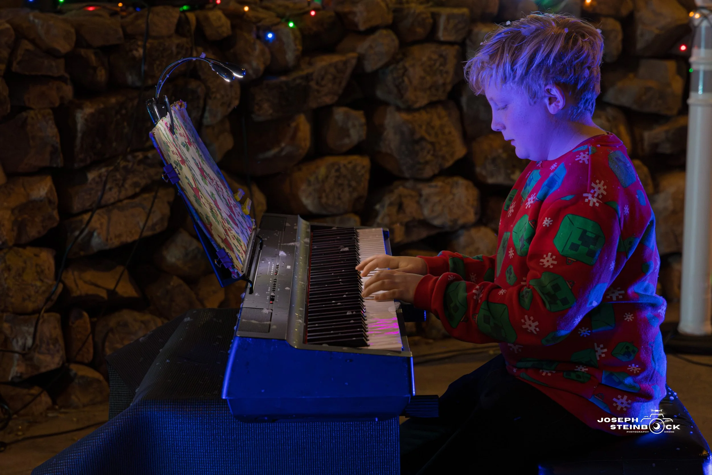 A young person playing an electric keyboard or piano, wearing a Christmas-themed sweater with green creeper faces and snowflakes, in front of a rocky wall with Christmas lights.