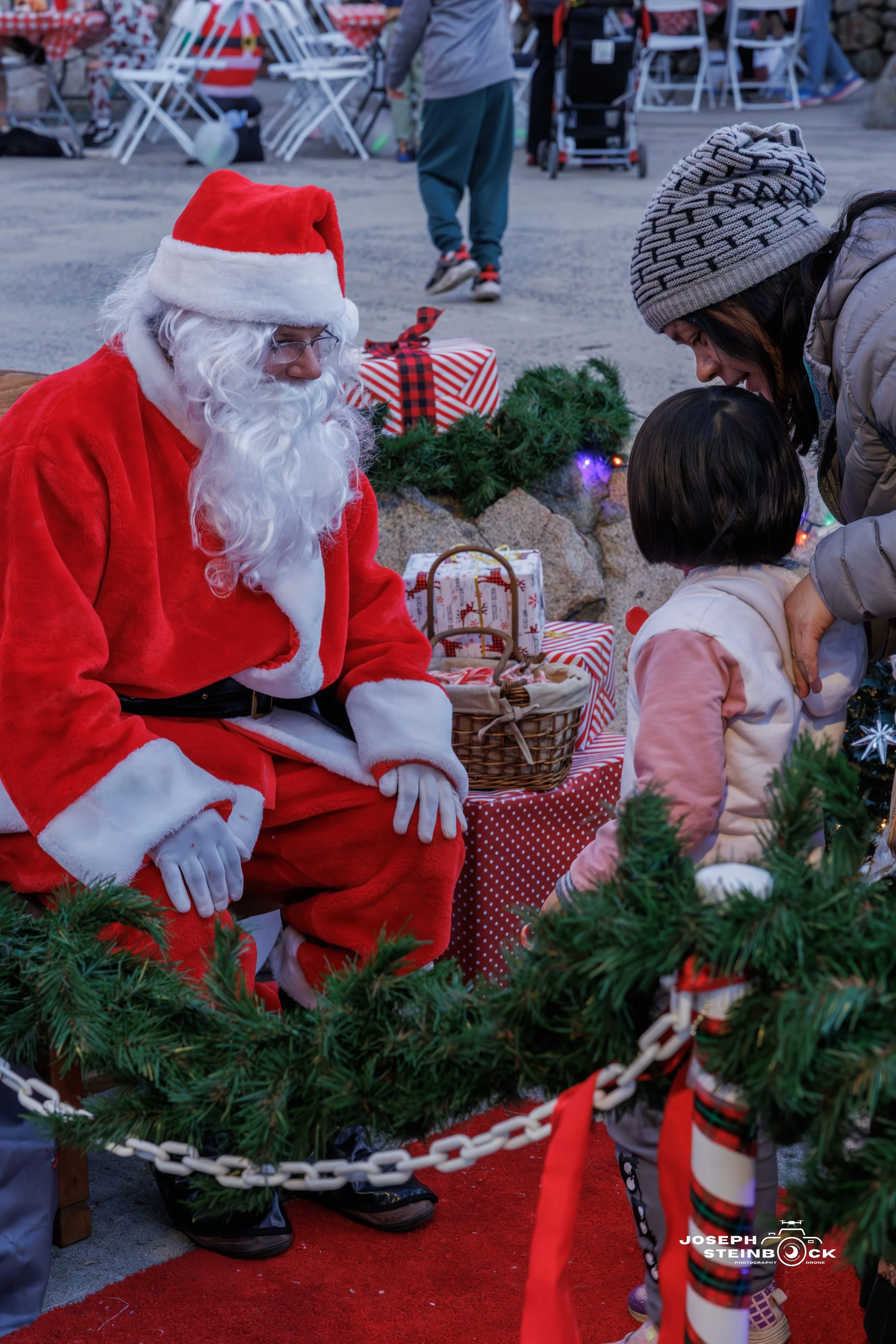 Santa Claus sitting on a bench, talking to a young girl and her mother during a Christmas event, surrounded by holiday decorations and gifts.