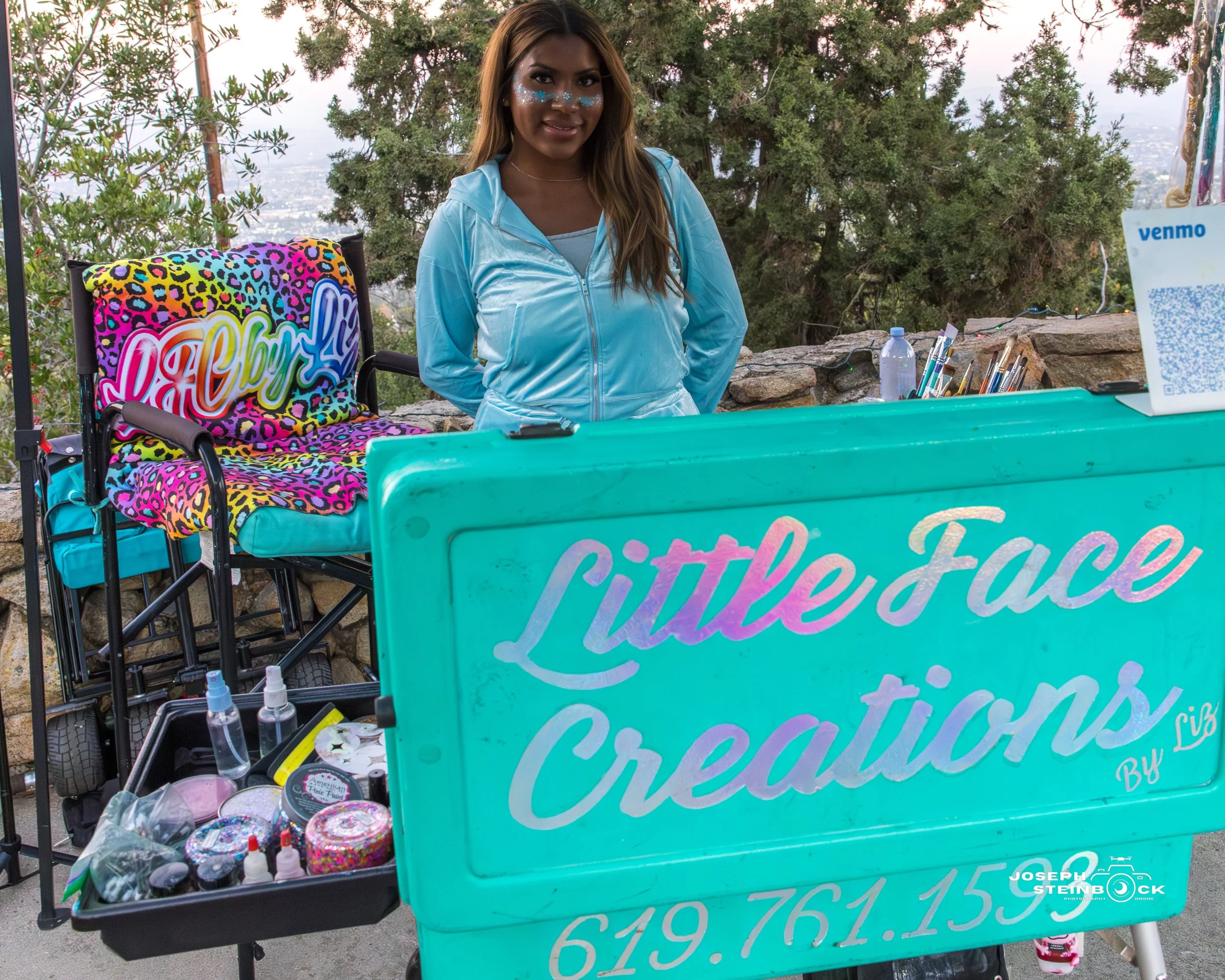 Woman standing behind a mint green cart with 'Little Face Creations' written on it in pastel colors, with makeup supplies and spray bottles, outdoors near a stone wall and trees.