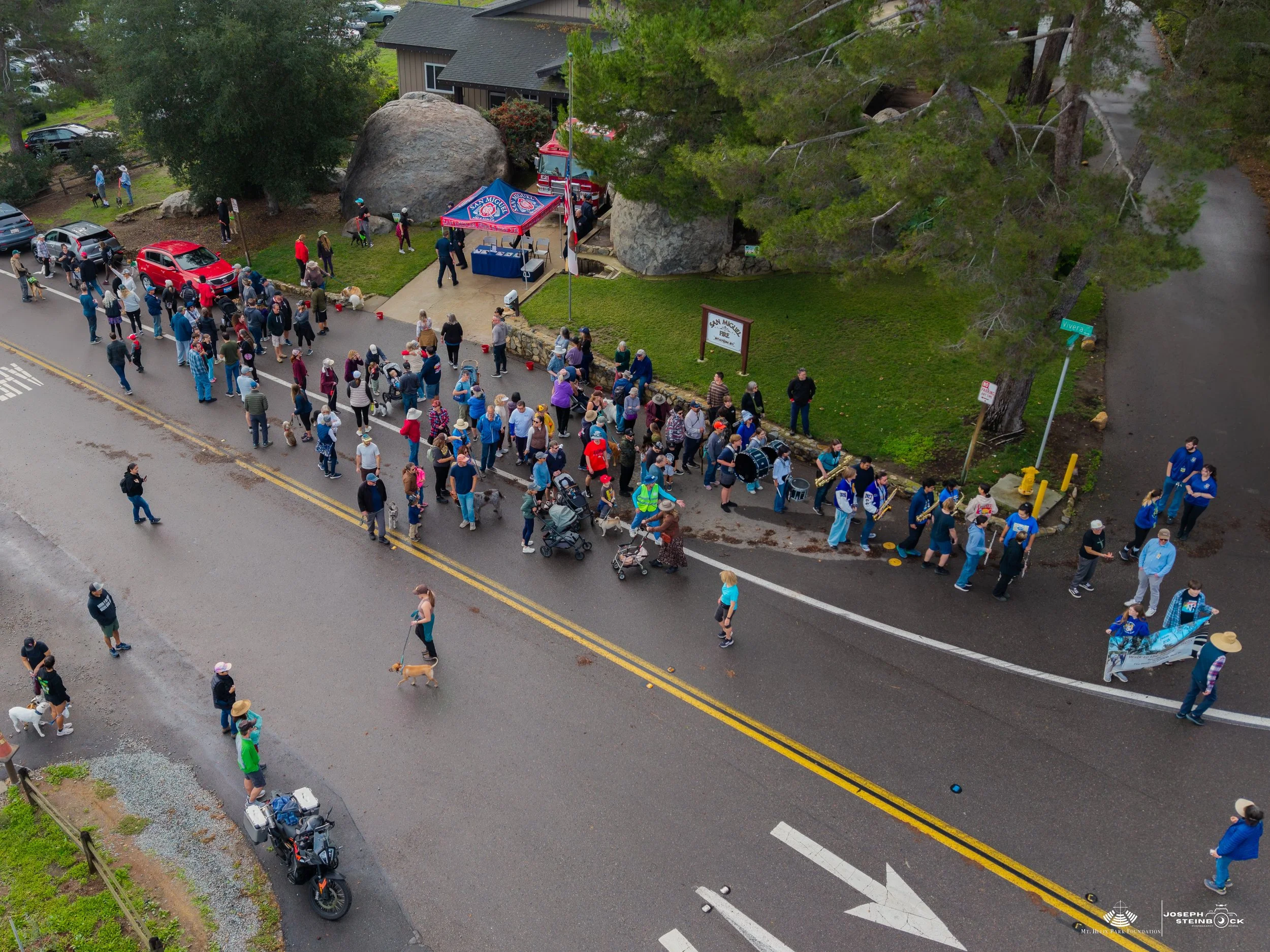 Aerial view of a large crowd lining the sidewalk and street outside a building with a sign that reads 'San Miguel Fire'. People are waiting in line, some holding umbrellas. Several fire trucks are parked near the building. Trees and parked cars are a