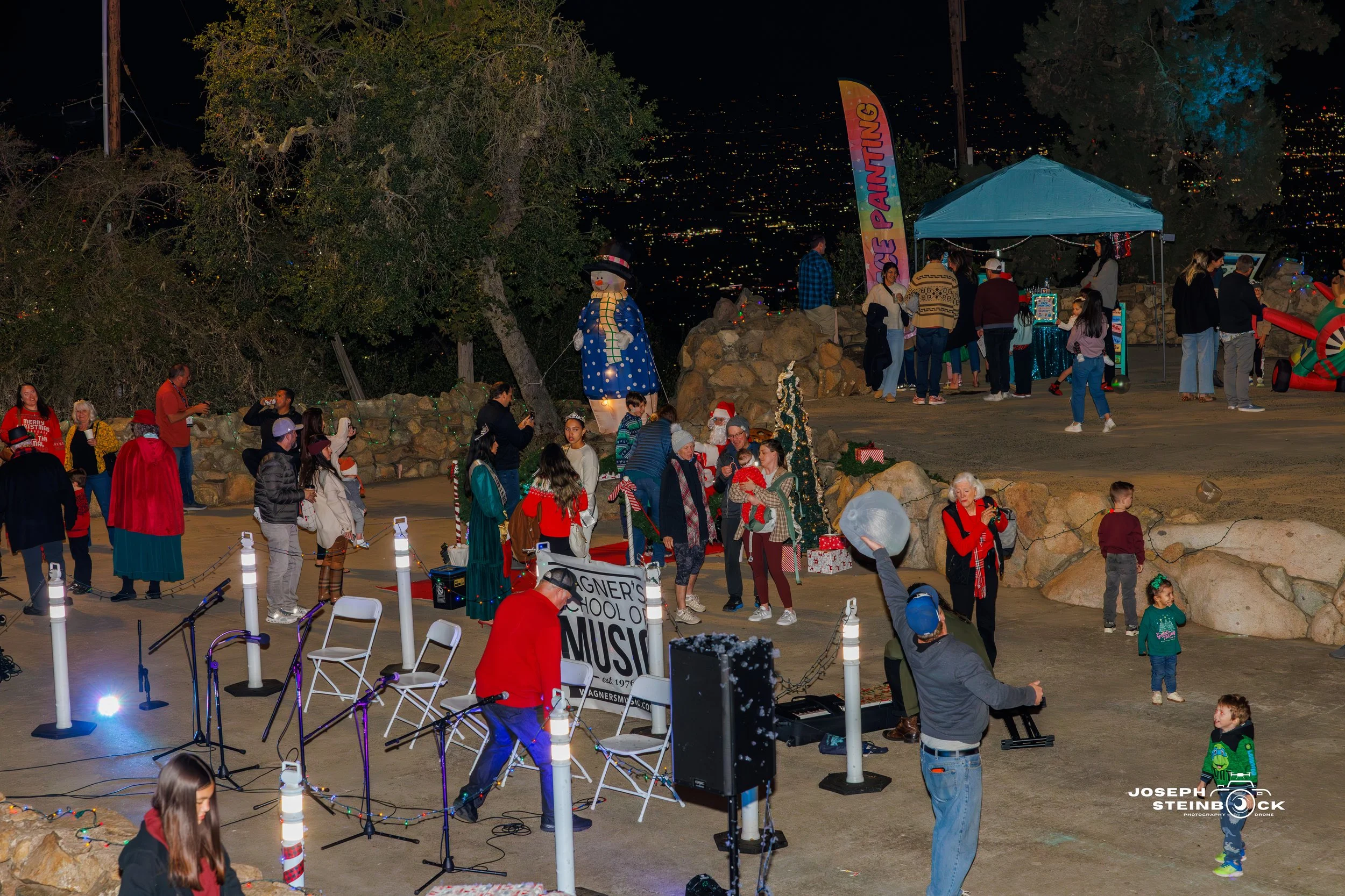 Outdoor night scene with people gathered at a festive event, including a musician performing with chairs and equipment, children playing, and holiday decorations. There is a snowman figure and a colorful banner that says 'Paining.'