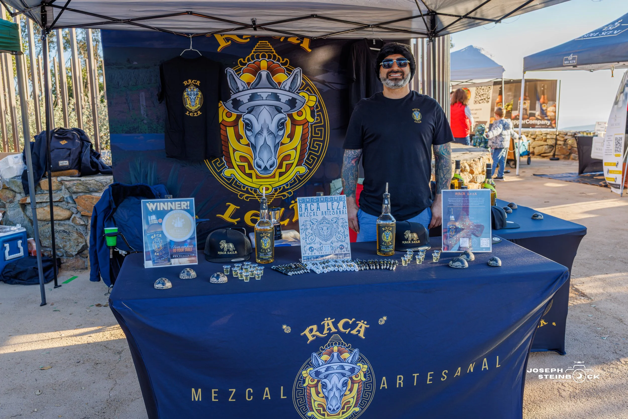 A man standing behind a table at an outdoor booth with a large logo of a donkey wearing a sombrero, displaying bottles of mezcal, shot glasses, hats, and promotional materials for Raca Leza Mezcal Artesanal.