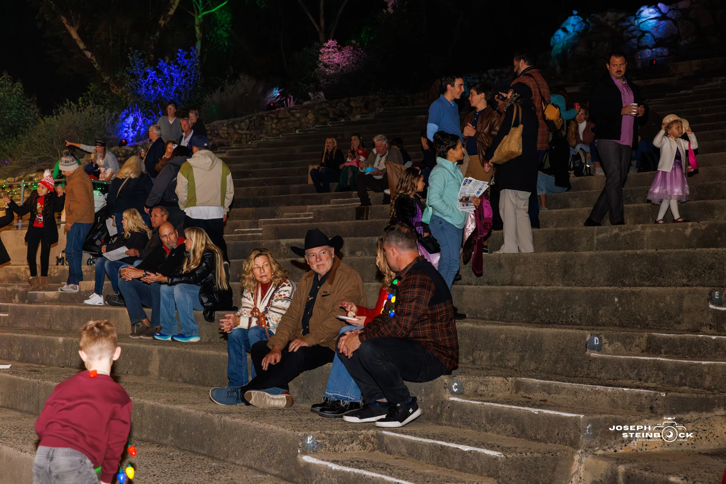 People sitting and standing on outdoor concrete staircase at night, some wearing festive or casual clothing, with colorful lights and trees in the background.