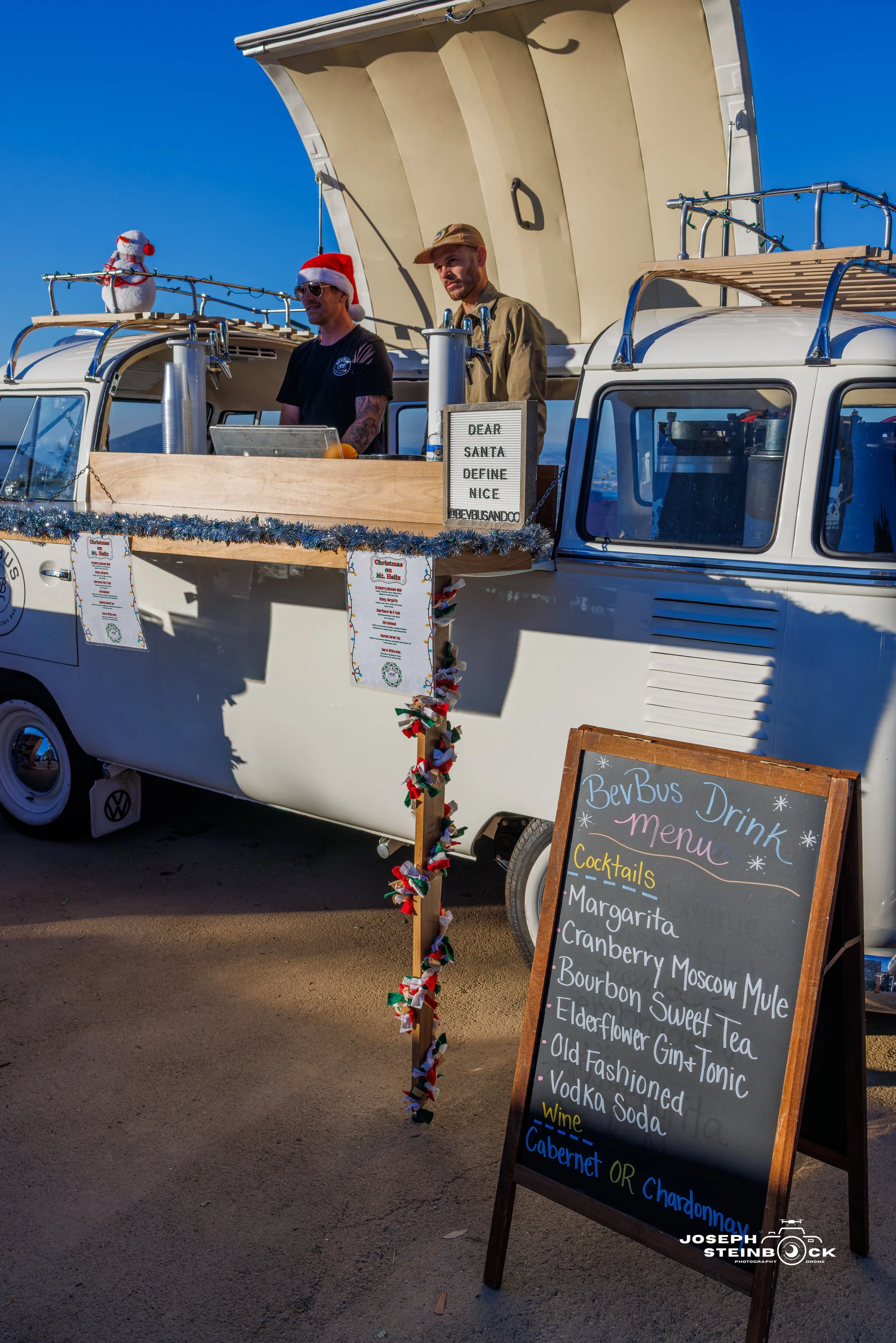 A vintage Volkswagen van decorated with Christmas decorations, serving as a mobile bar with a menu sign listing cocktails and wine options. Two men stand behind the counter, one wearing a Santa hat, with a small snowman on the roof. A chalkboard sign