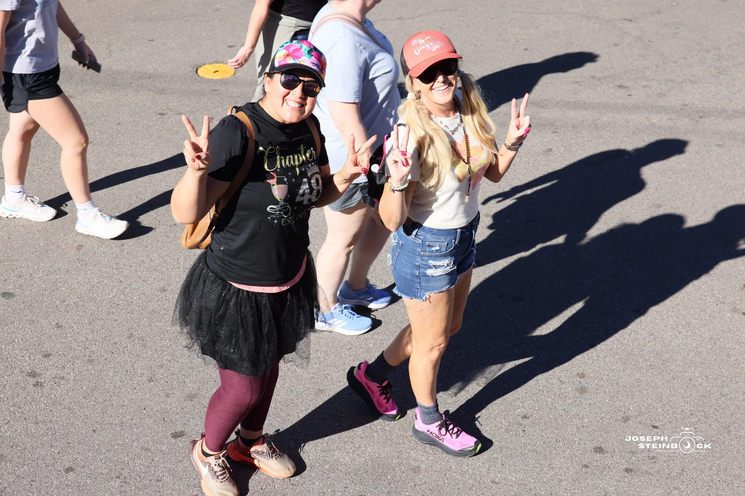 Two women walking outdoors, smiling, and making peace signs with their hands. One wears a black T-shirt, a black tutu, maroon tights, and sneakers. The other wears a white T-shirt, denim shorts, and pink running shoes. They are shaded on the ground, 