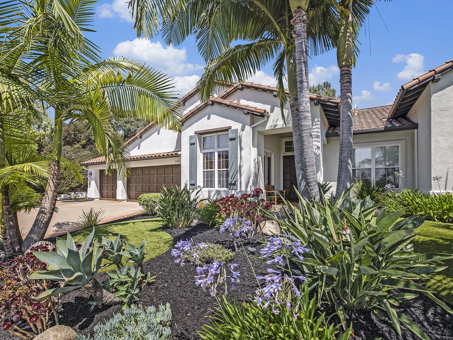 Front yard of a suburban house with lush green plants, purple flowers, tall palm trees, and a brown garage door, under a blue sky with scattered white clouds.