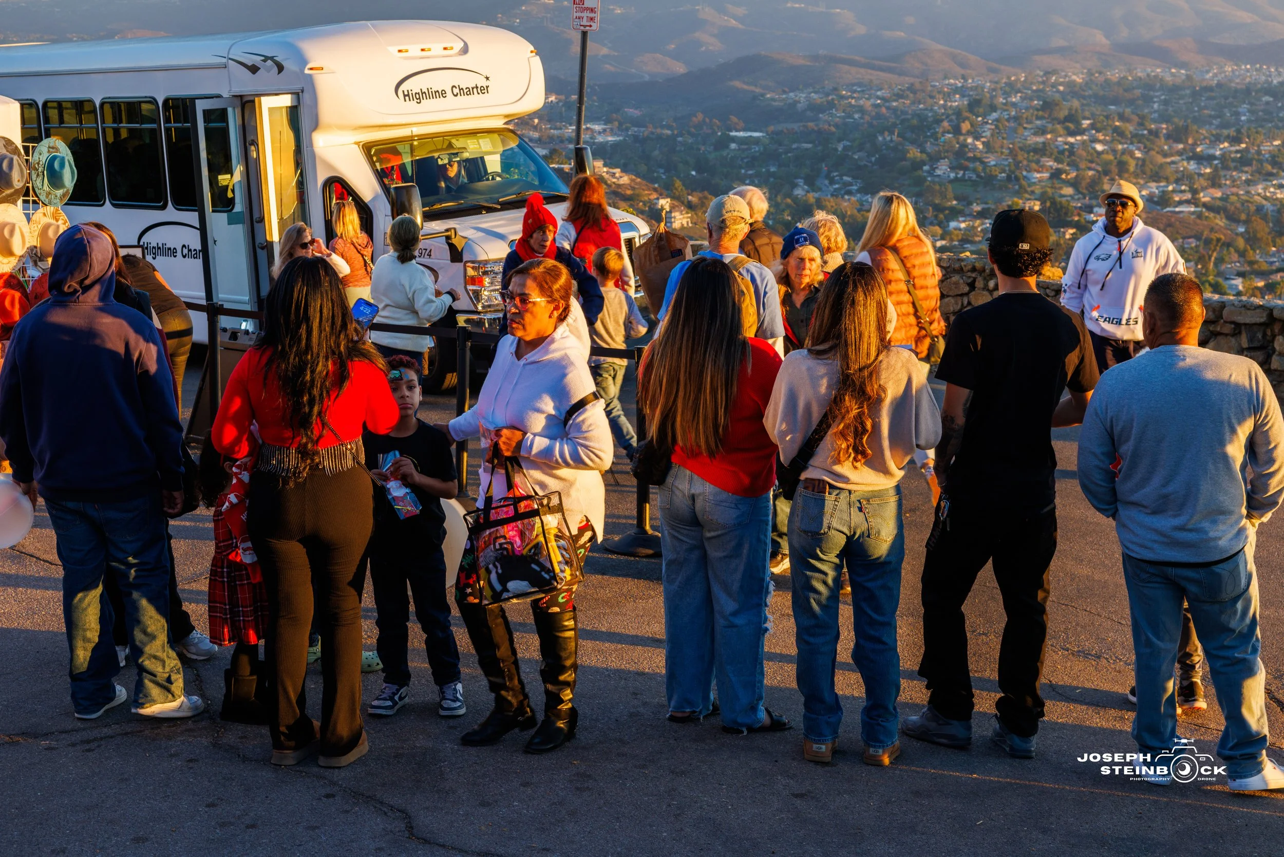 People gathered outdoors near a sightseeing bus, overlooking a cityscape with hills in the background during sunset.