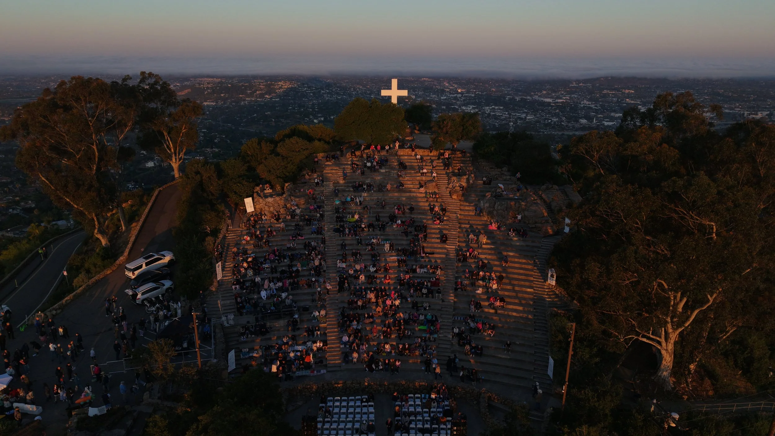 A large outdoor gathering with many people sitting on tiered steps at a park during sunset, with a large white cross at the top of the hill and city skyline in the background.