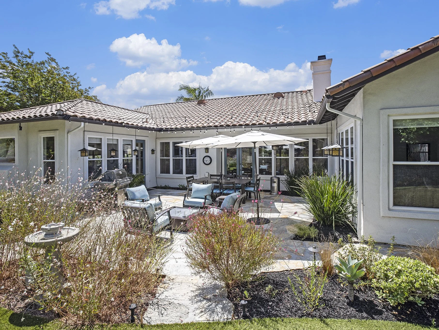 Backyard patio with outdoor furniture, umbrellas, garden, and house with tiled roof under blue sky.