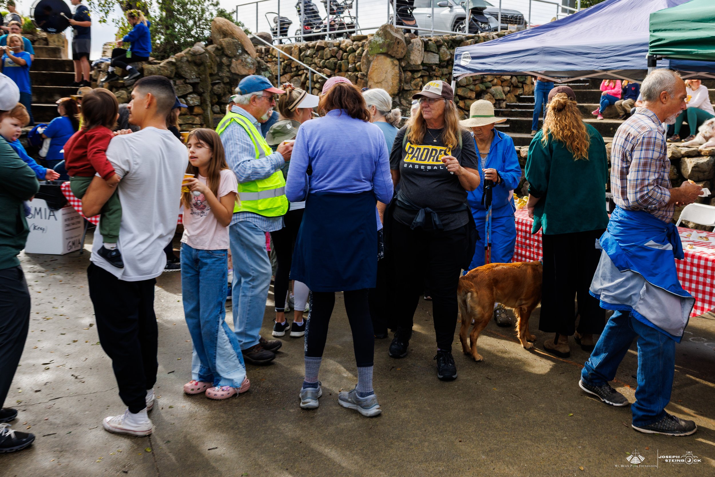People gathered at an outdoor community event, with some standing under tents and others near a table. There are children and adults, some engaging in conversations, and a dog on a leash. The scene is lively with a stone staircase in the background a