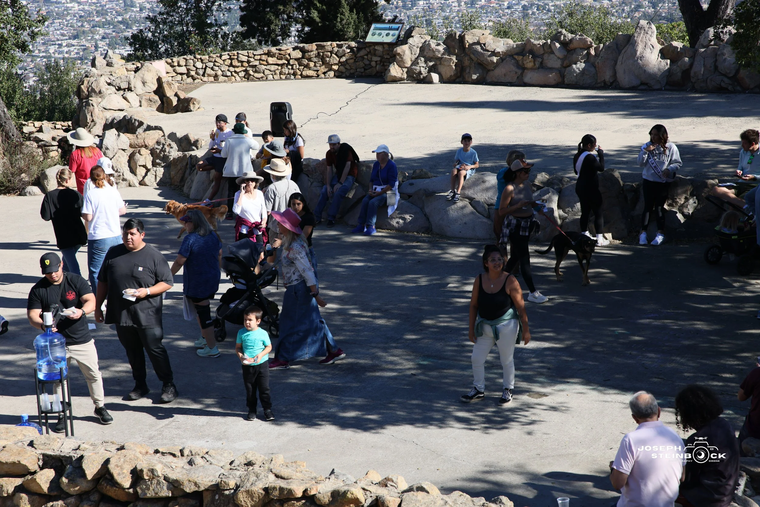 People gathered at an outdoor event in a park-like setting with rock formations, some sitting on rocks and others walking around, while a water dispenser is visible in the foreground. Shade from trees creates patterns on the ground.