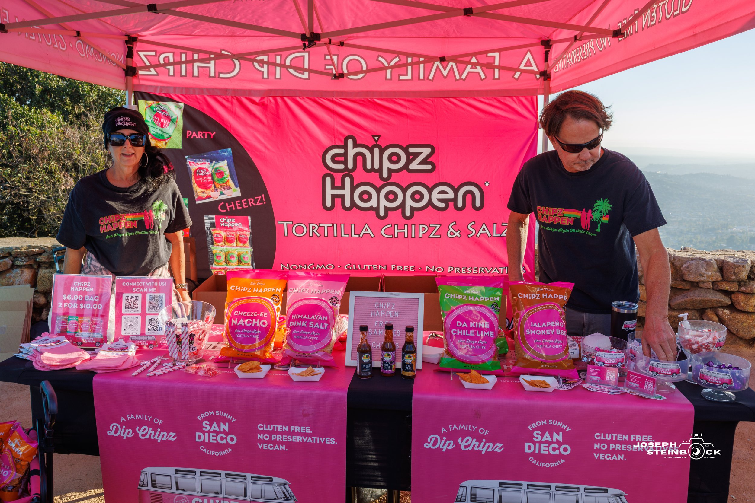 Two people standing behind a pink promotional table for Chipz Happen at an outdoor event, with food bags and bottles on display.