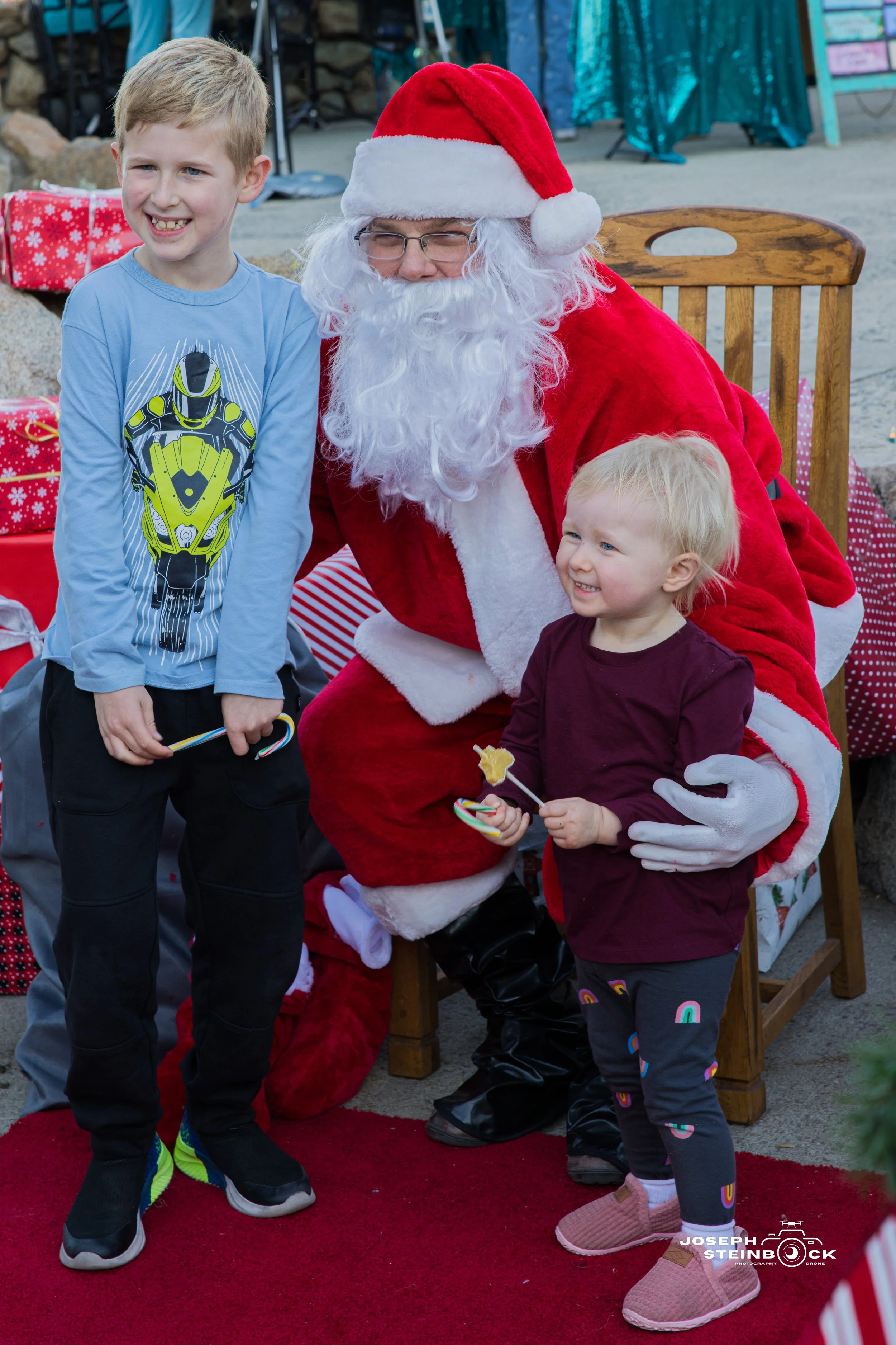 A man dressed as Santa Claus sitting in a chair with a young boy and girl. The boy, standing to Santa's left, is smiling and holding a candy cane. The girl, standing to Santa's right, is smiling, holding a lollipop, and wearing pink shoes. Santa has 