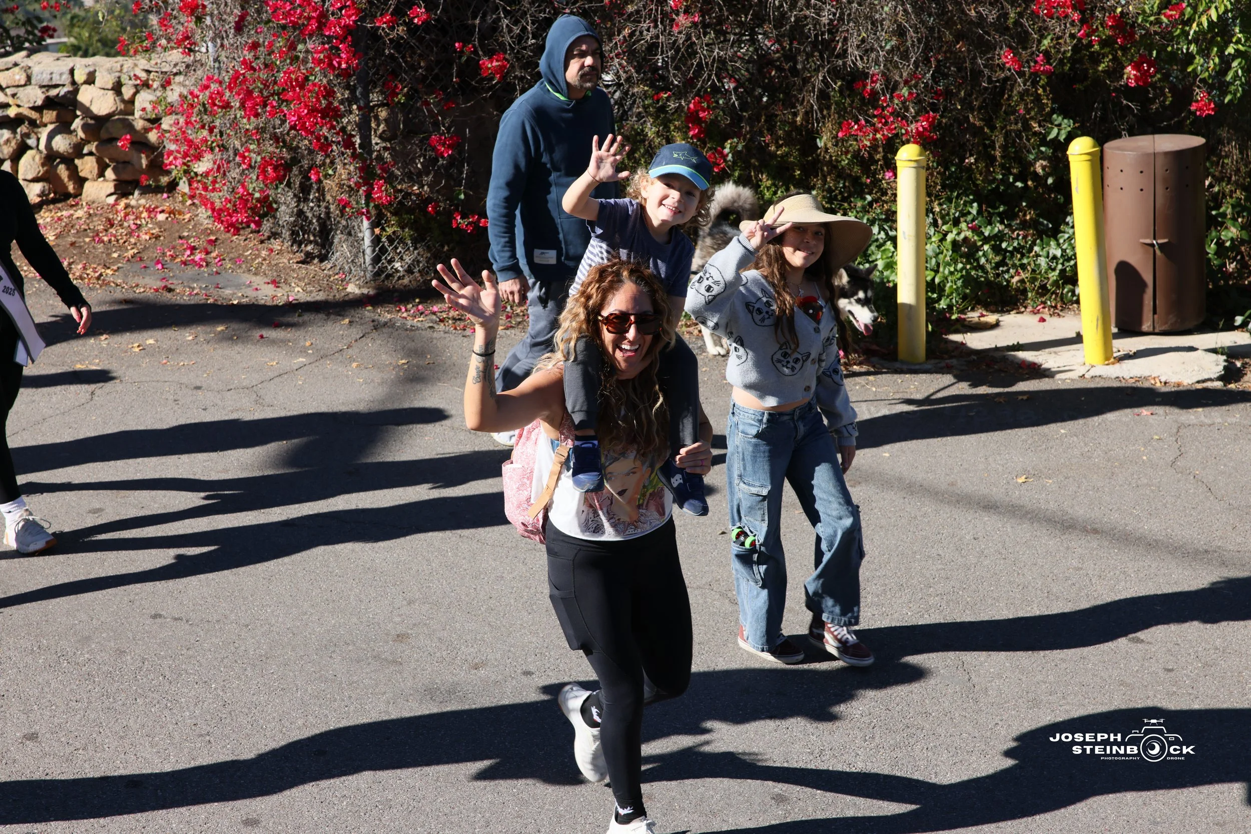 A group of people, including women and children, walking outdoors on a paved area with shrubbery and flowers in the background. The group appears to be cheerful, with some waving and smiling at the camera.
