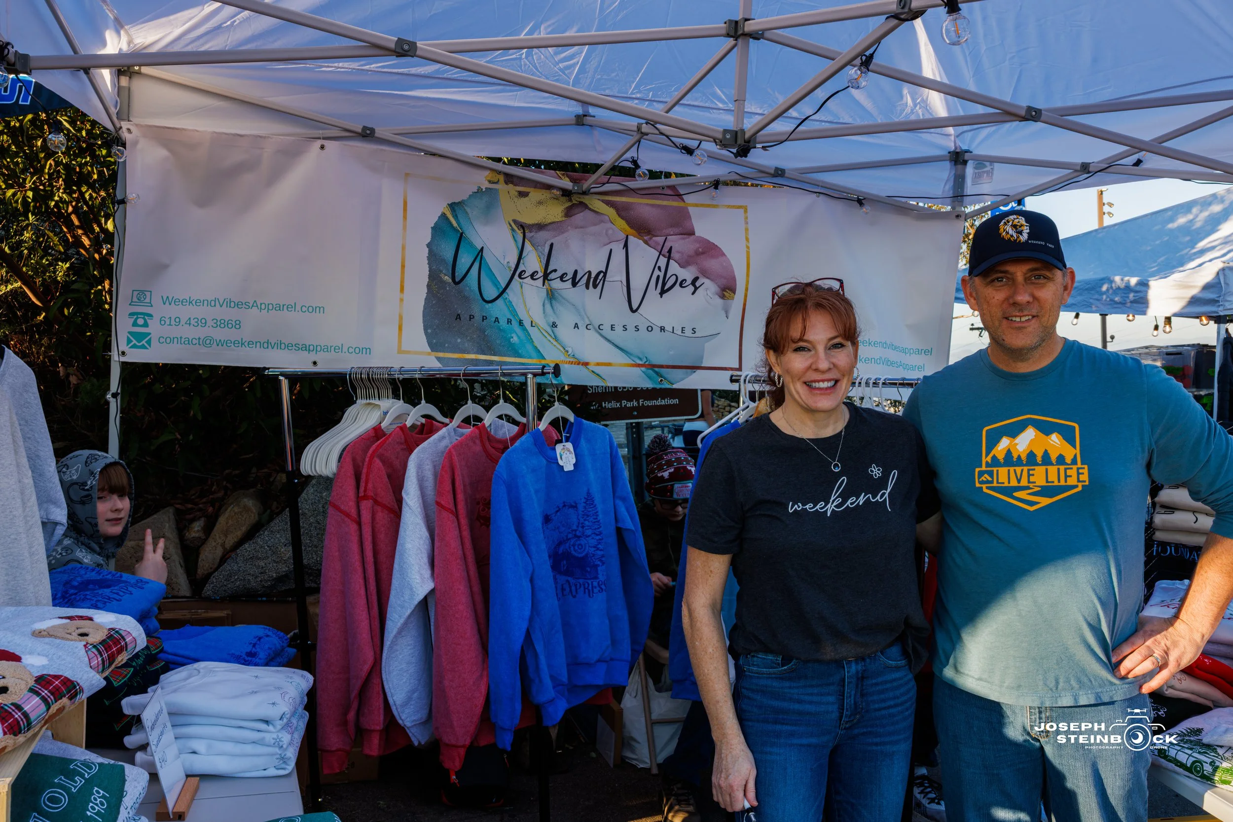 Two people standing in front of a booth selling clothing at an outdoor market, with a sign that reads "Weekend Vibes Apparel & Accessories." There are colorful sweatshirts hanging on a rack behind them, and other clothing items on a table to the left