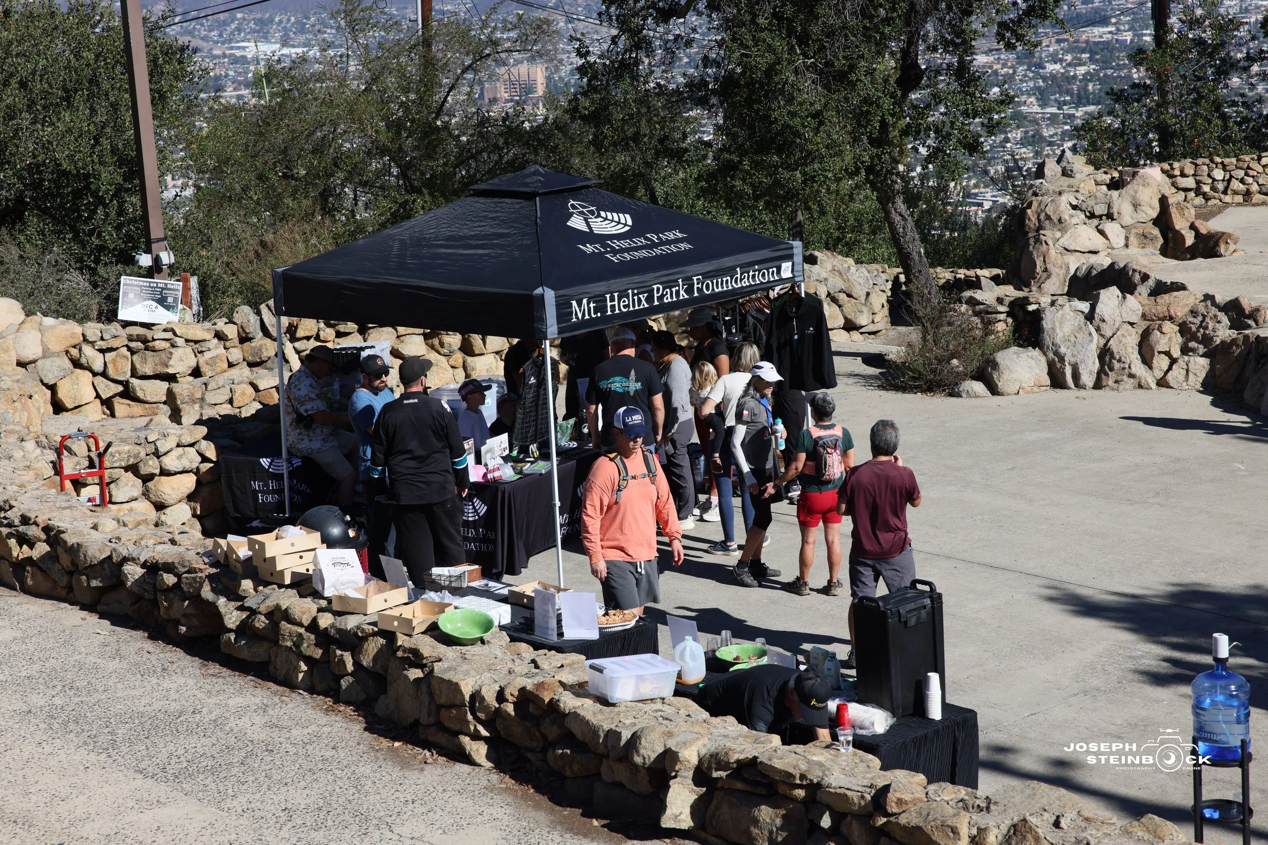 A group of people gathered under a black tent with the words "Mt. Helix Park Foundation" written on it, at an outdoor event at Mt. Helix Park. The area is surrounded by rocky borders, and some tables with food and supplies are set up.