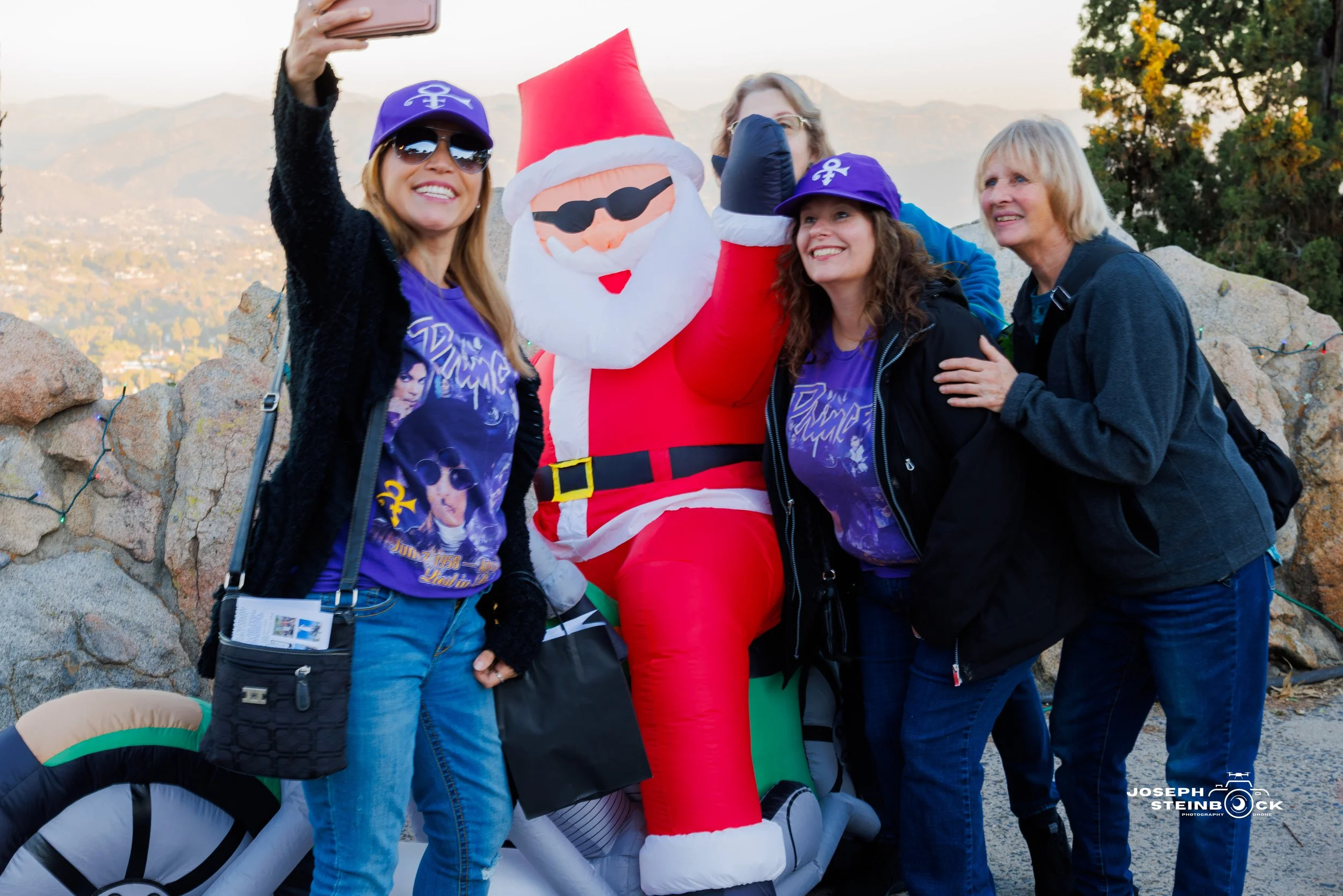 Four women taking a selfie with a person in a Santa Claus inflatable costume outdoors during daytime.