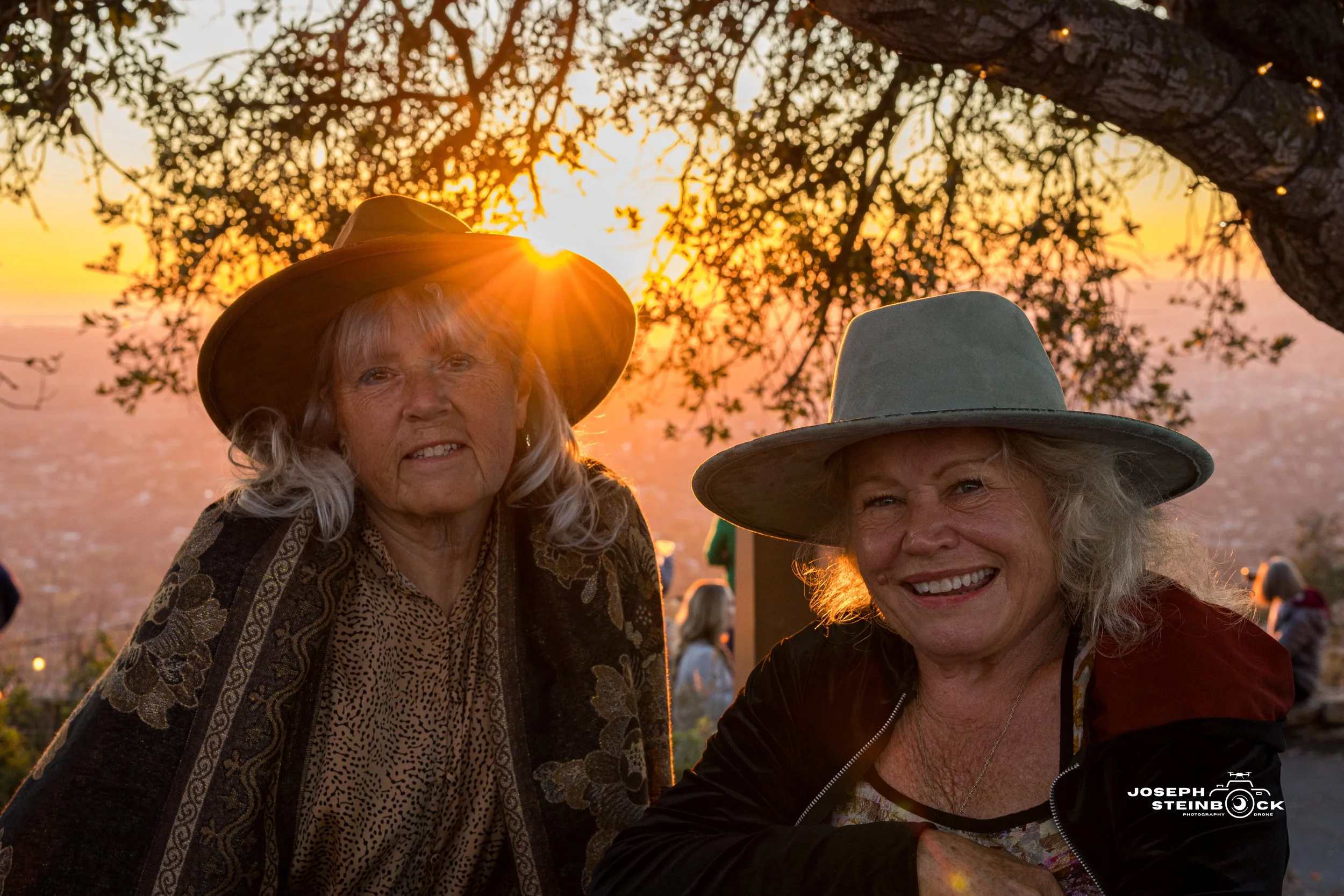 Two elderly women with light skin and gray hair wearing wide-brimmed hats, smiling outdoors at sunset under a tree, with other people in the background.