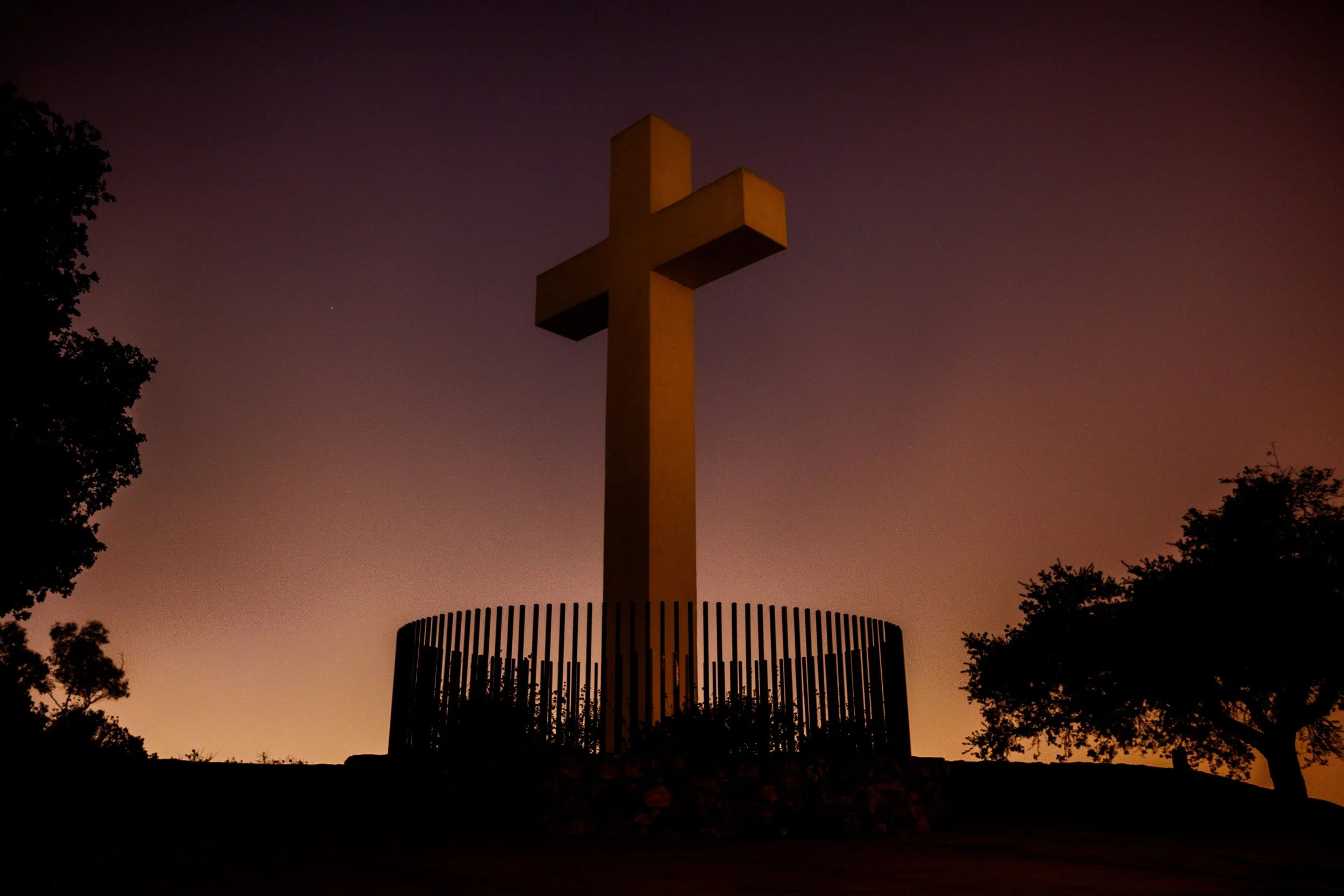 Large cross monument silhouetted against a colorful dusk or dawn sky, surrounded by a circular fence and trees.