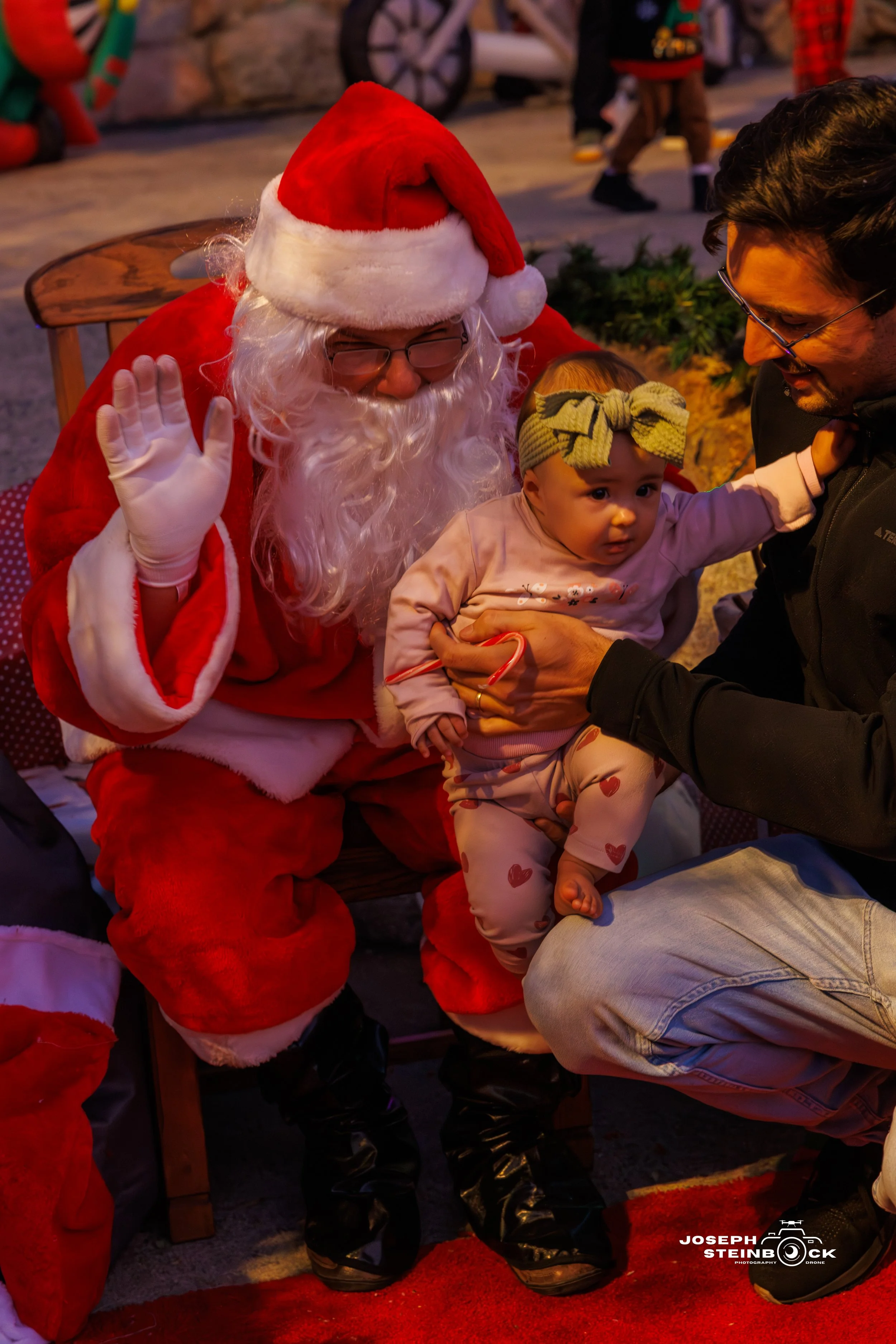 Santa Claus in traditional red outfit, sitting on a wooden bench, waving hello, with a baby girl sitting on a man's lap who is holding her. The baby girl is wearing pajamas with hearts and a green headband with a bow. The scene appears to be festive,