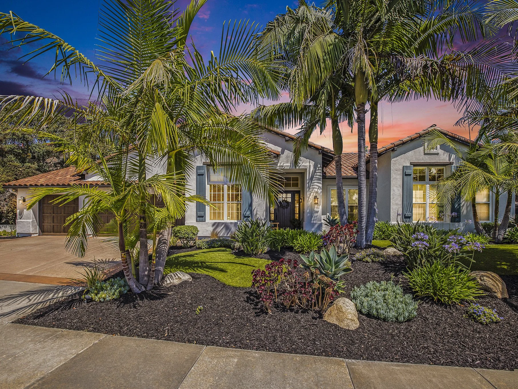 A house with a well-maintained front yard featuring palm trees, flowering plants, and a paved driveway at sunset.