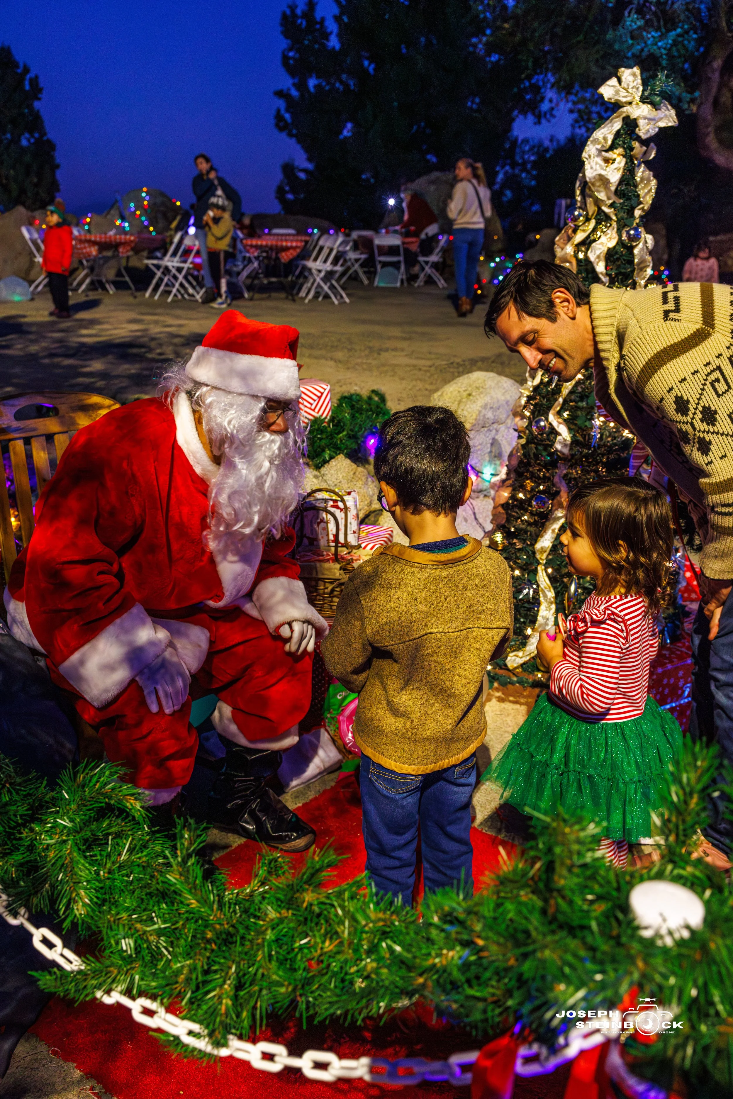 Santa Claus sitting outside with two children, a man, and a woman, near a decorated Christmas tree at night.