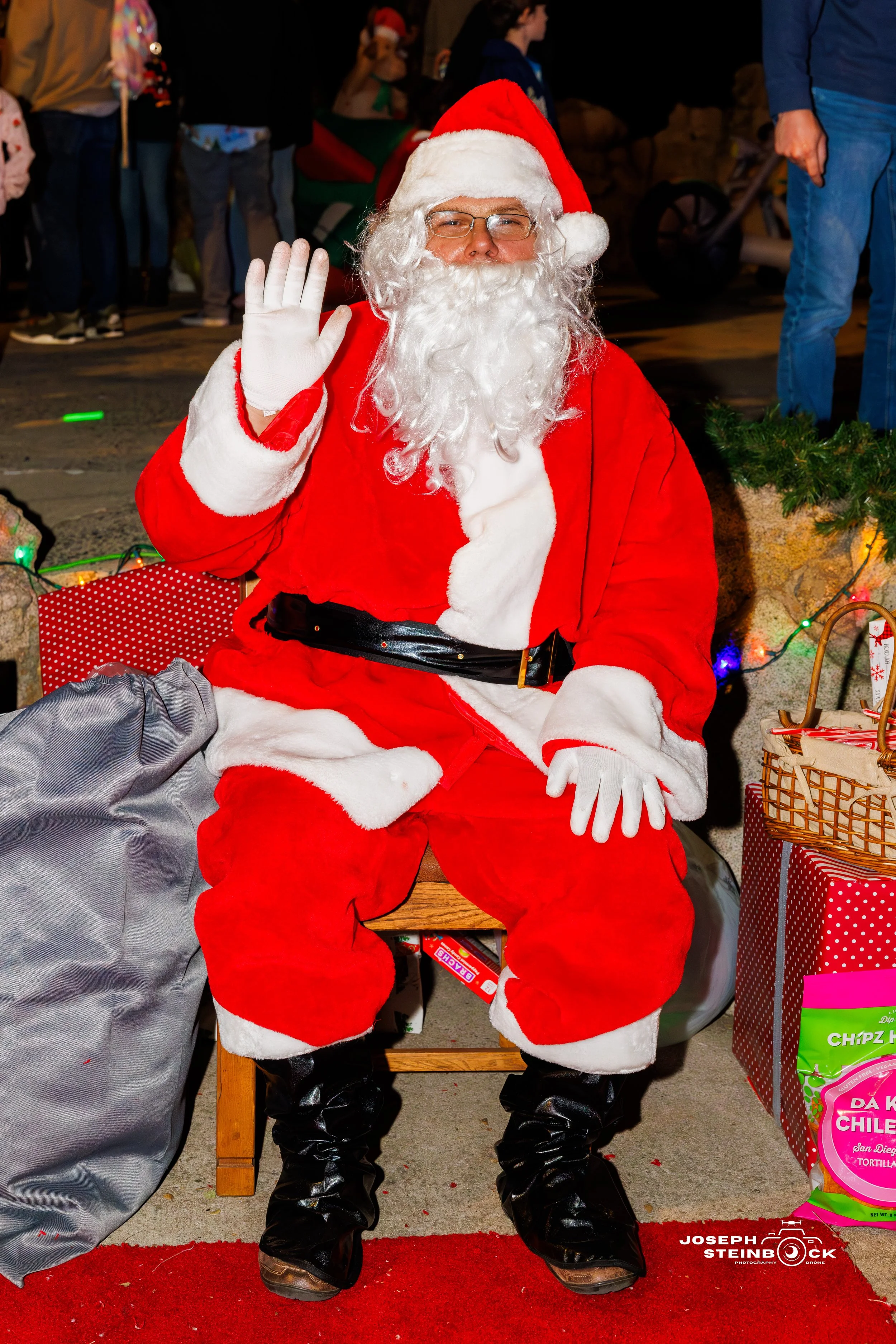 Man dressed as Santa Claus sitting on a bench, waving, at a Christmas event.