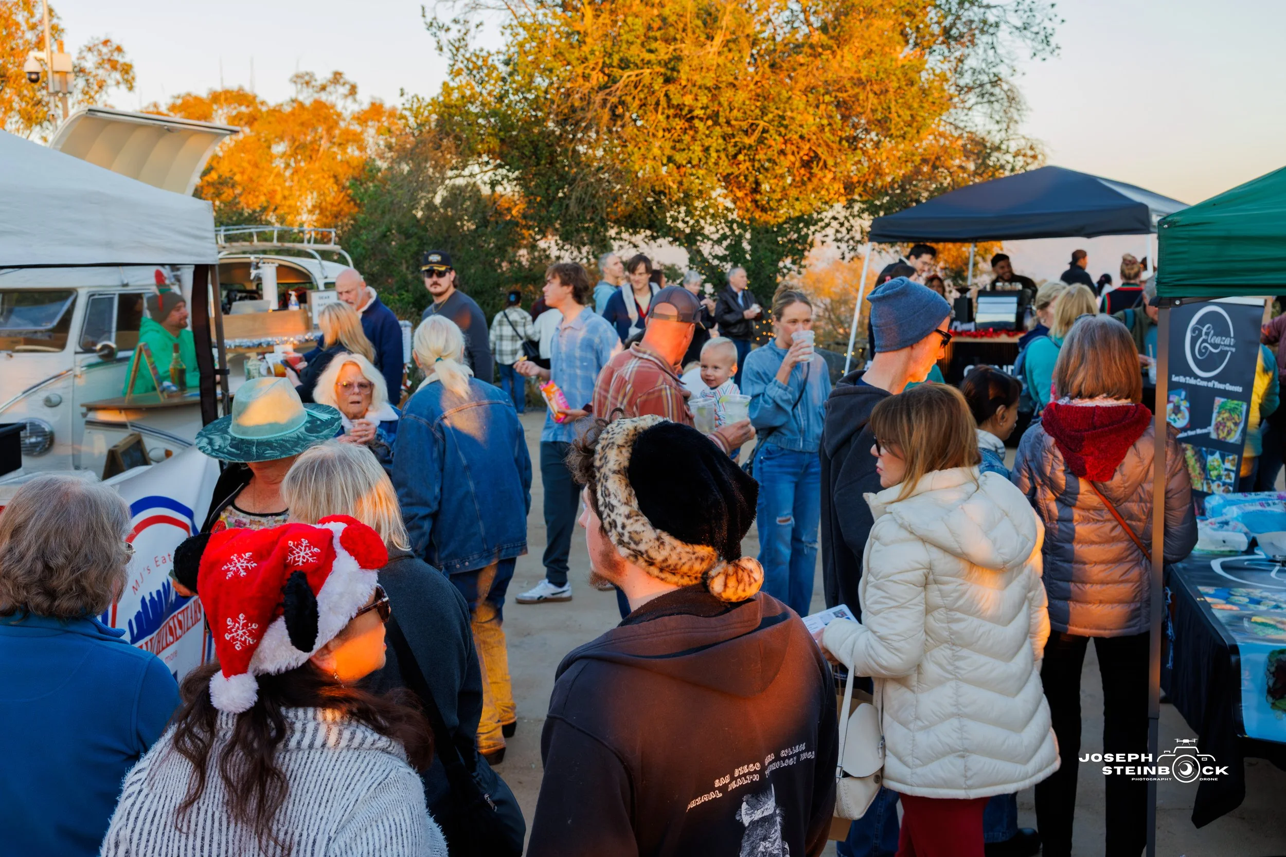 Crowd of people at an outdoor market or festival during fall, with colorful autumn trees in the background.