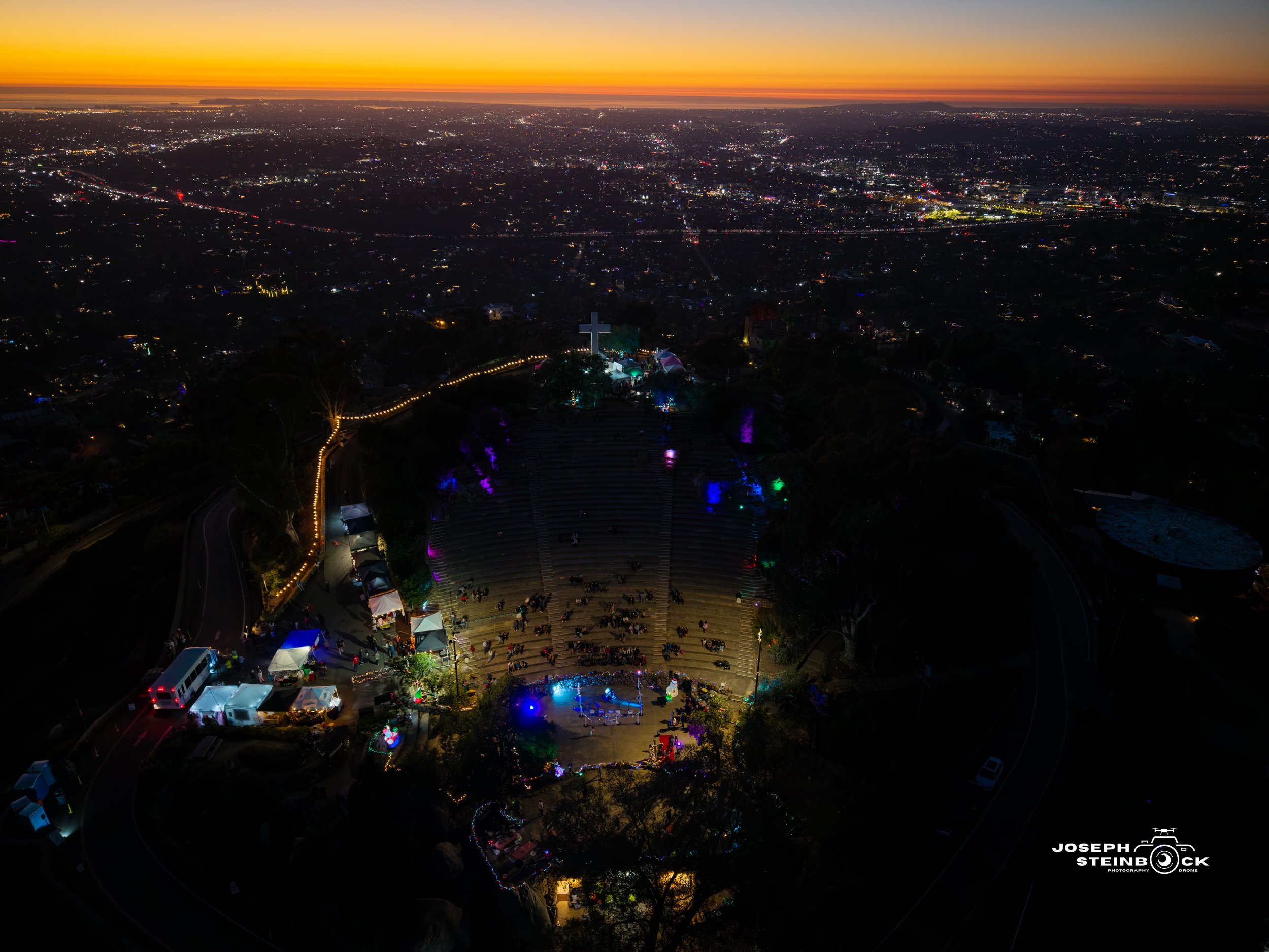 An aerial night view of an outdoor gathering at a park with illuminated tents, a stage, and a cross, overlooking a cityscape with many lights and a sunset sky in the background.