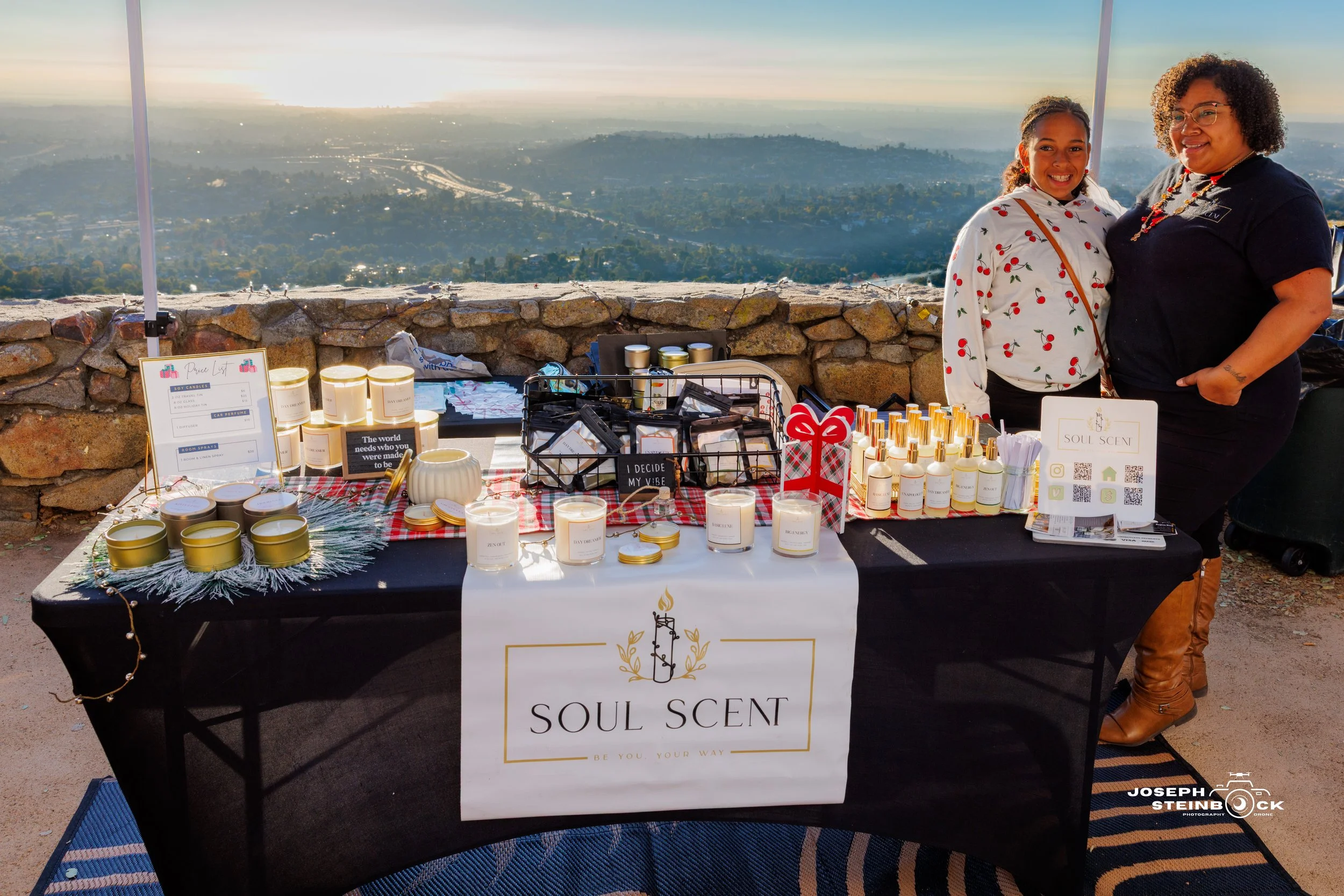 A woman and a girl stand beside a table with candles and products labeled 'Soul Scent' at an outdoor event on a scenic overlook with a city and hills in the background.
