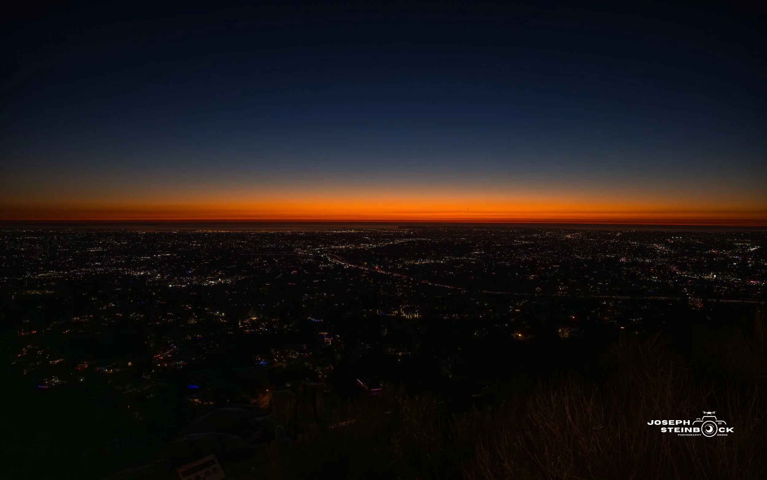 Sunset view over a cityscape with a darkening sky, faint stars, and illuminated city lights.