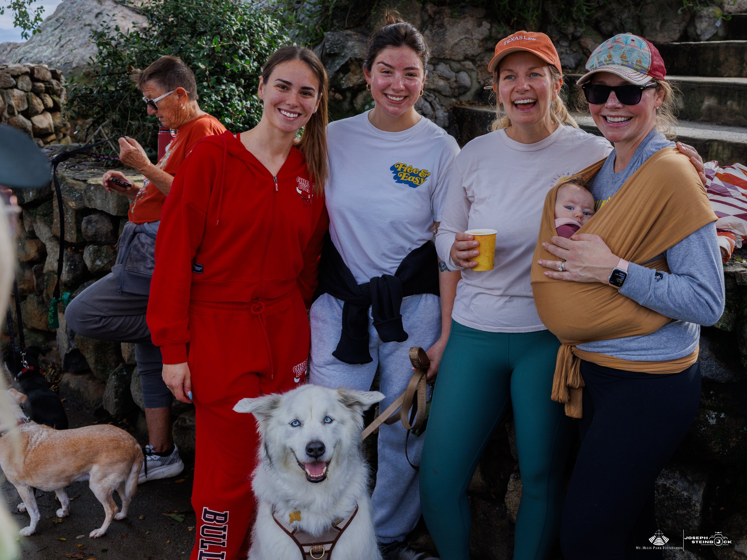 Group of five women and two dogs, one on a leash and one in a baby sling, outdoor scene with rock steps.
