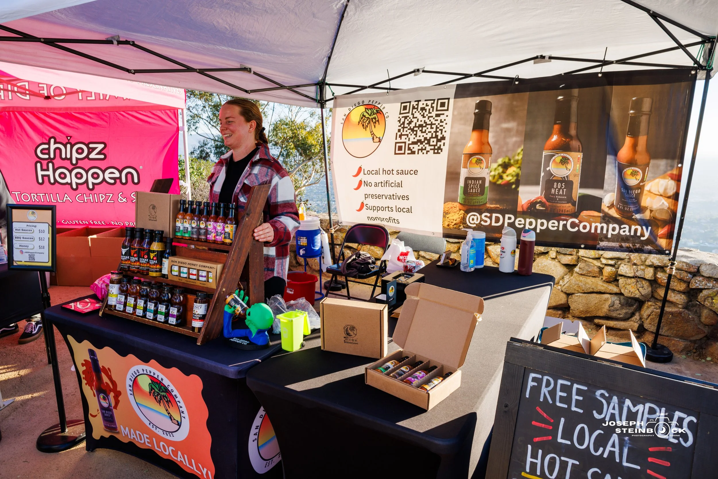 A woman standing at a booth selling hot sauce and honey products at an outdoor market. The booth has banners promoting local and natural products, with jars of honey and bottles of hot sauce displayed on the table.