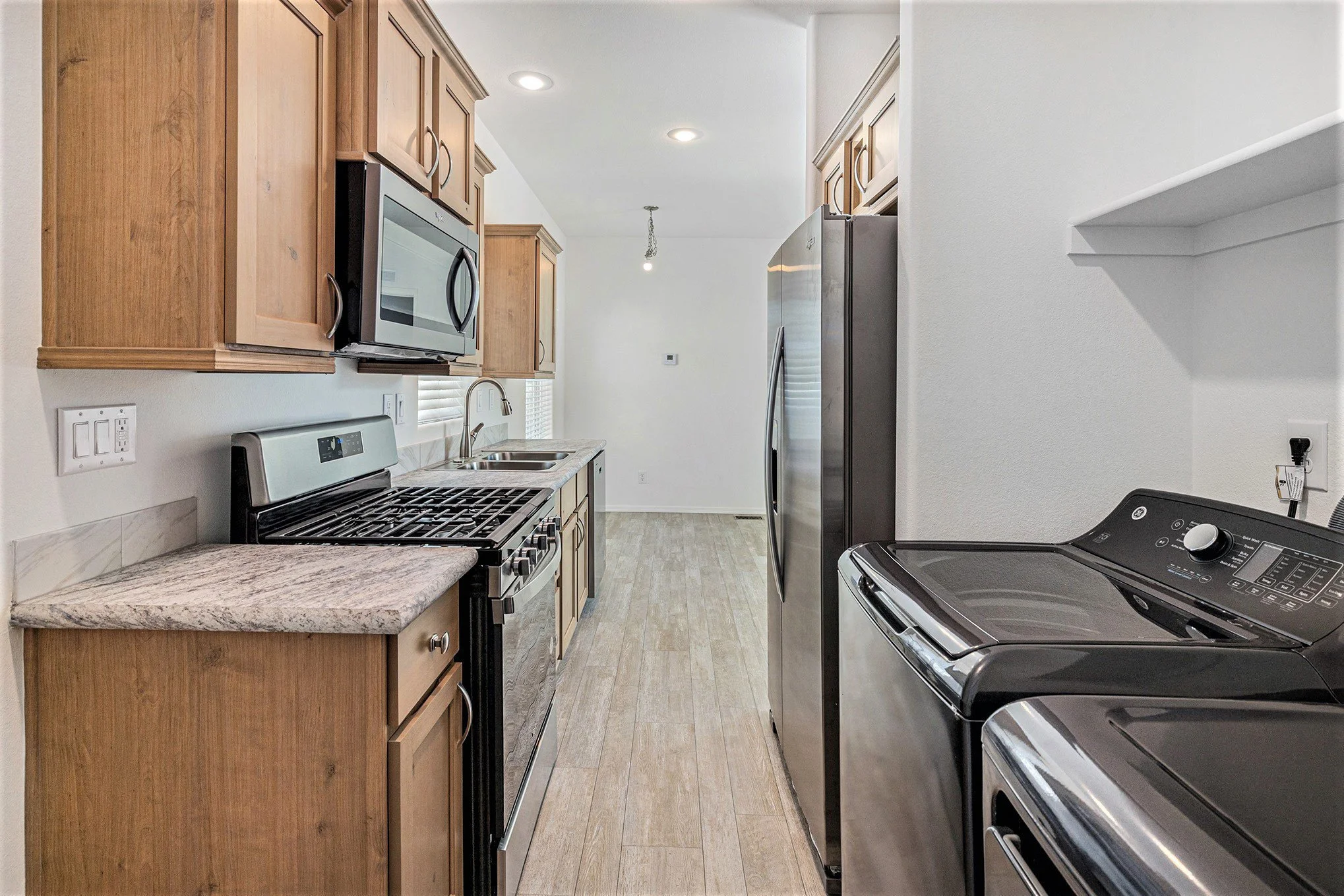 Kitchen with wooden cabinets, stainless steel appliances including a microwave, fridge, stove, and a washing machine, with a double sink under a window and recessed ceiling lights.