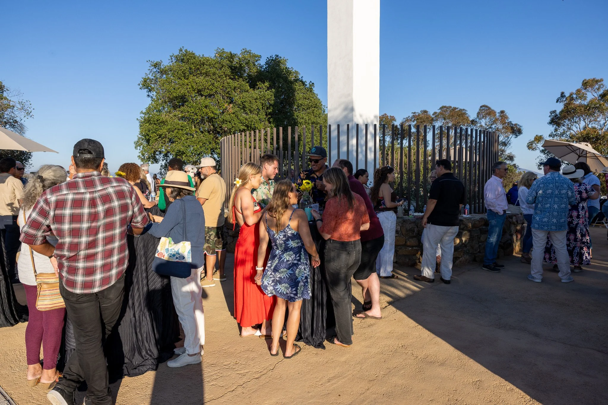 Group of people gathered outdoors near a large monument, socializing and enjoying a sunny day, with trees in the background.