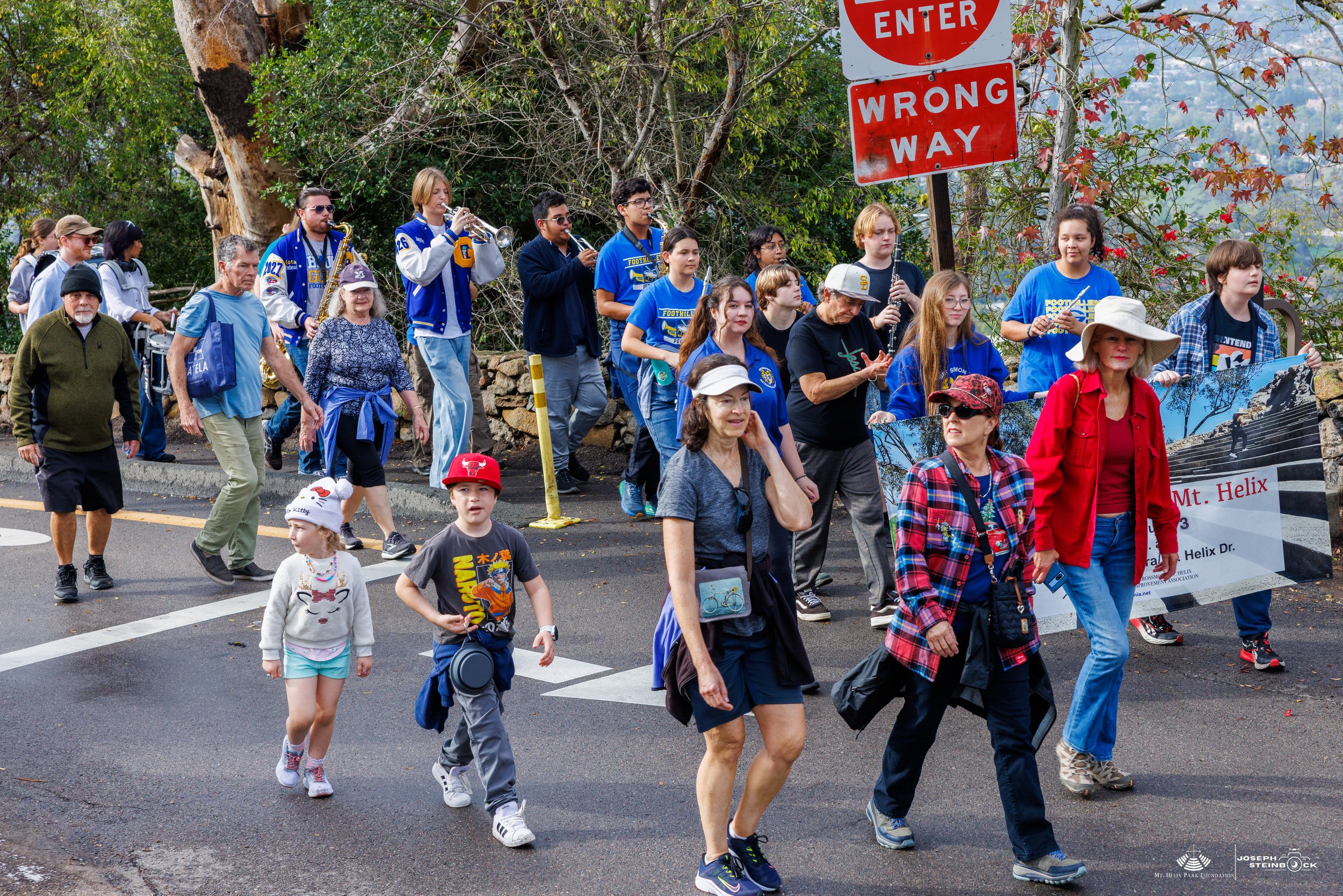 A diverse group of people, including children, adults, and the elderly, walking on a street during a parade or event. Several people are holding signs and a large banner advertising Mount Helix. Some are wearing casual and colorful clothing, and ther