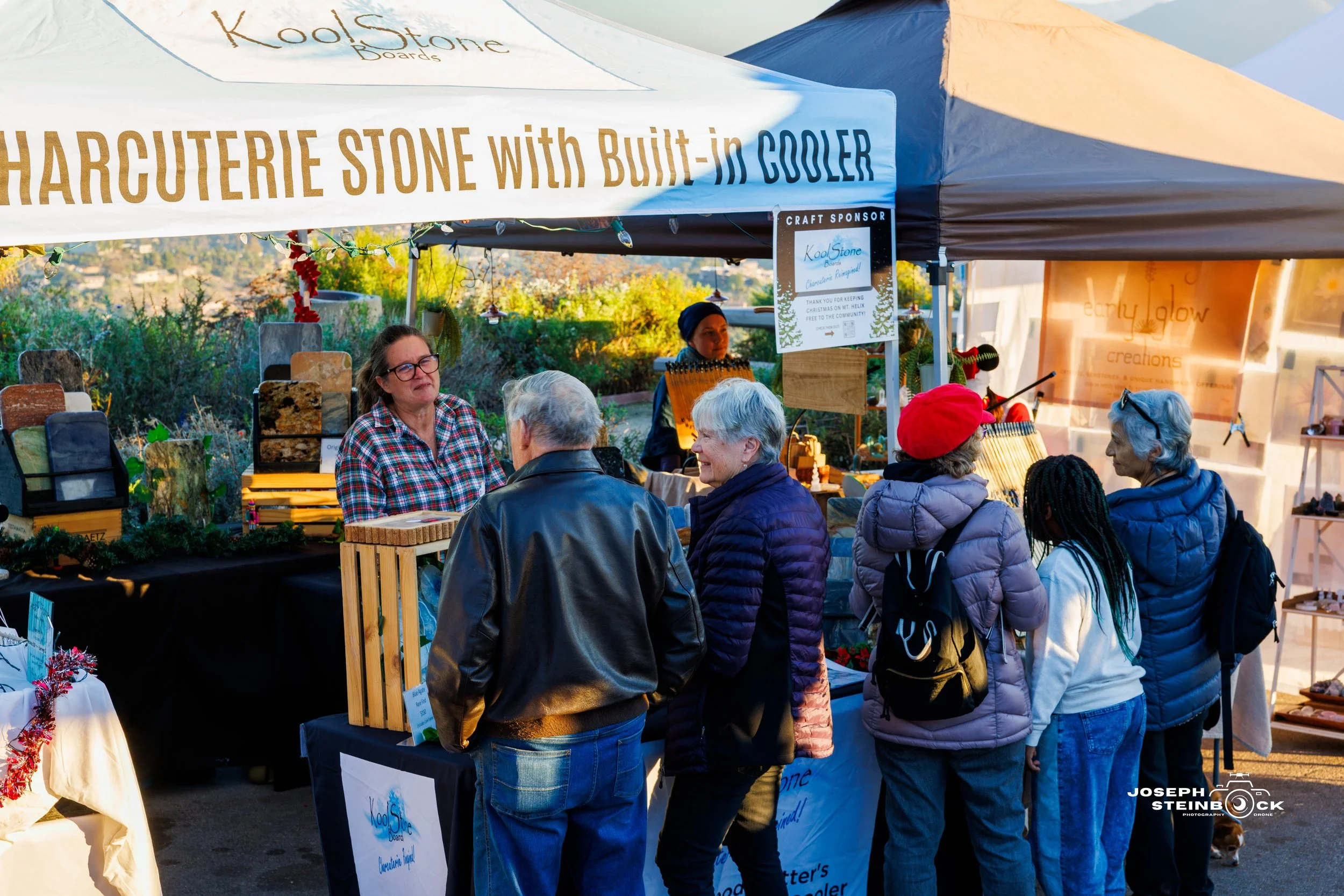 People browsing a market stall with stone boards and a built-in cooler, under a white canopy with a banner that reads 'KoolStone.' The vendor is behind the stall, and there are trees and hills in the background.