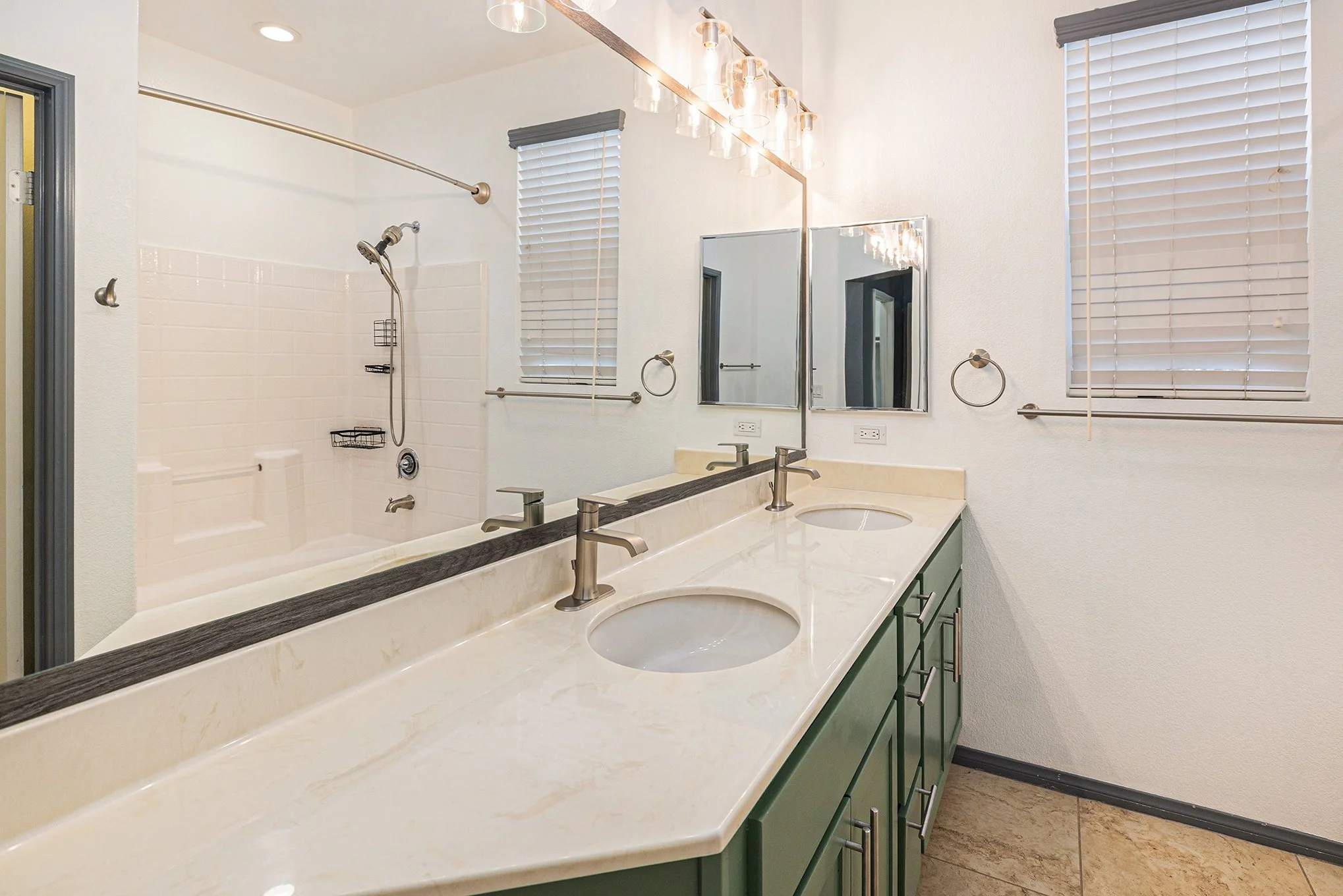 Bathroom with double sink vanity, large mirror, wall-mounted lights, window with blinds, and a walk-in shower with white tile walls.