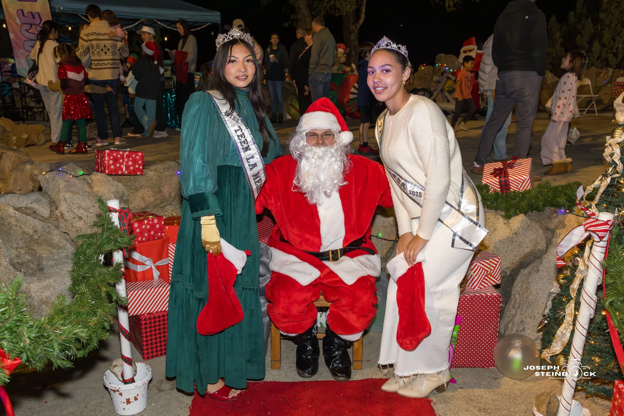 Two young women wearing sashes and crowns stand next to Santa Claus at a Christmas event. They are holding red stockings and surrounded by wrapped gifts, festive decorations, and a holiday setting with other people in the background.