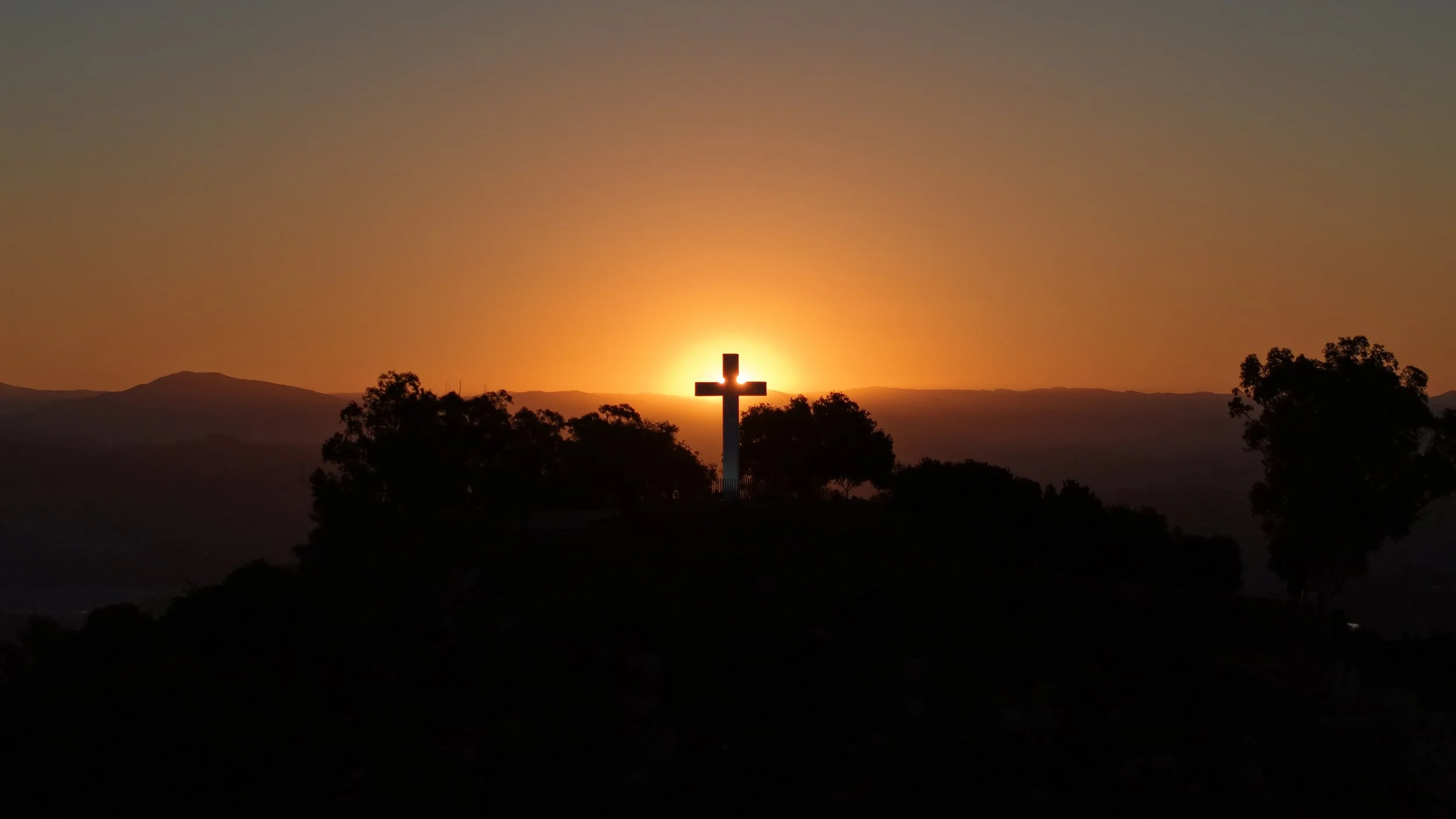 A silhouette of a Christian cross on a hilltop at sunset, with the sun setting behind it and trees in the foreground.