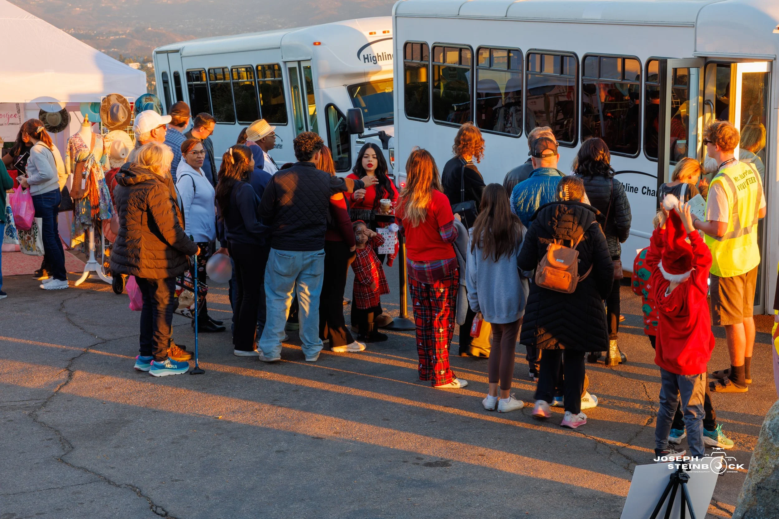 People waiting in line to board a small shuttle bus during a holiday event; some are wearing Christmas sweaters and Santa hats.