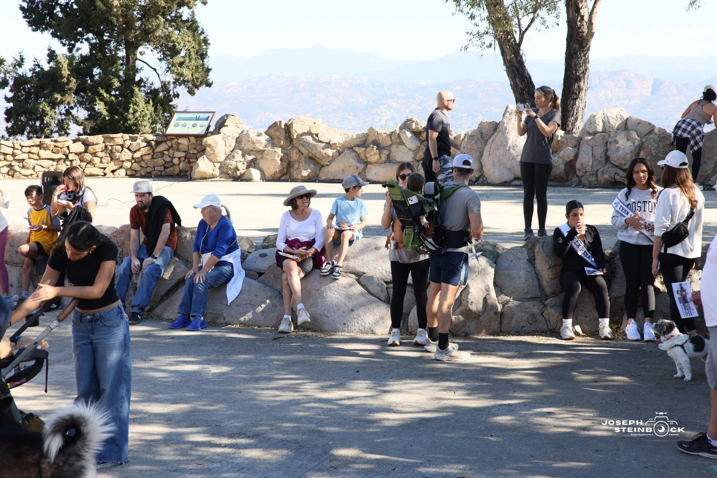 A group of people, including children and adults, sitting and standing on a rocky area with trees and a stone wall. Some are taking photos or talking, with mountains and a blue sky in the background.