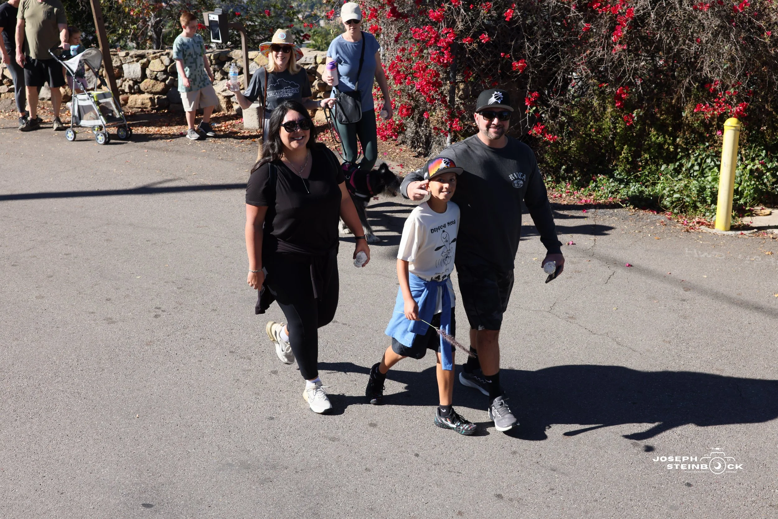 A group of six people, including two children, walking on a paved path outdoors on a sunny day. They are smiling and carrying water bottles. In the background, there are three other people, one with a stroller and another holding a water bottle, near