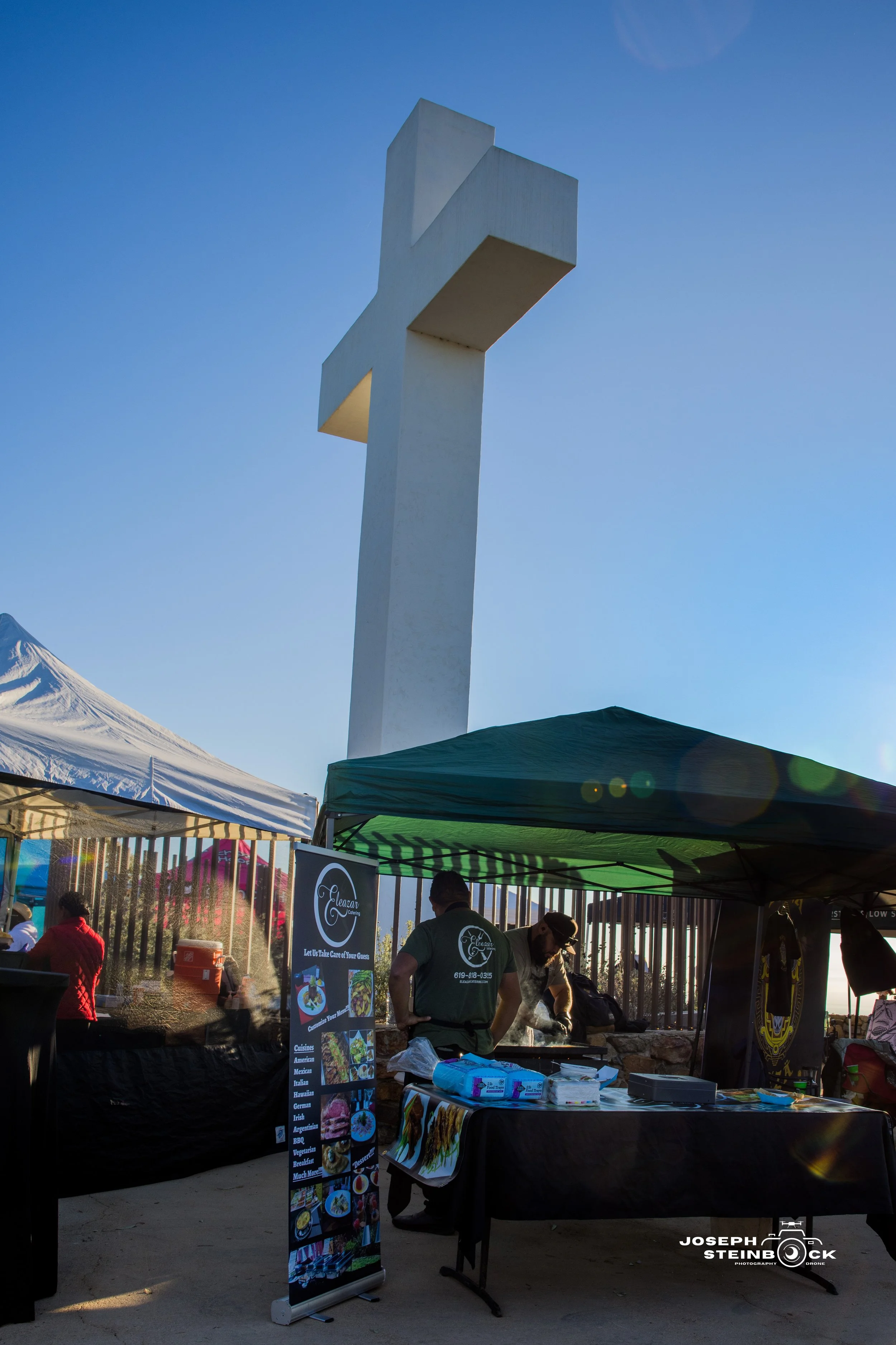 Outdoor market stall with food vendors under tents, with a large white cross monument in the background.