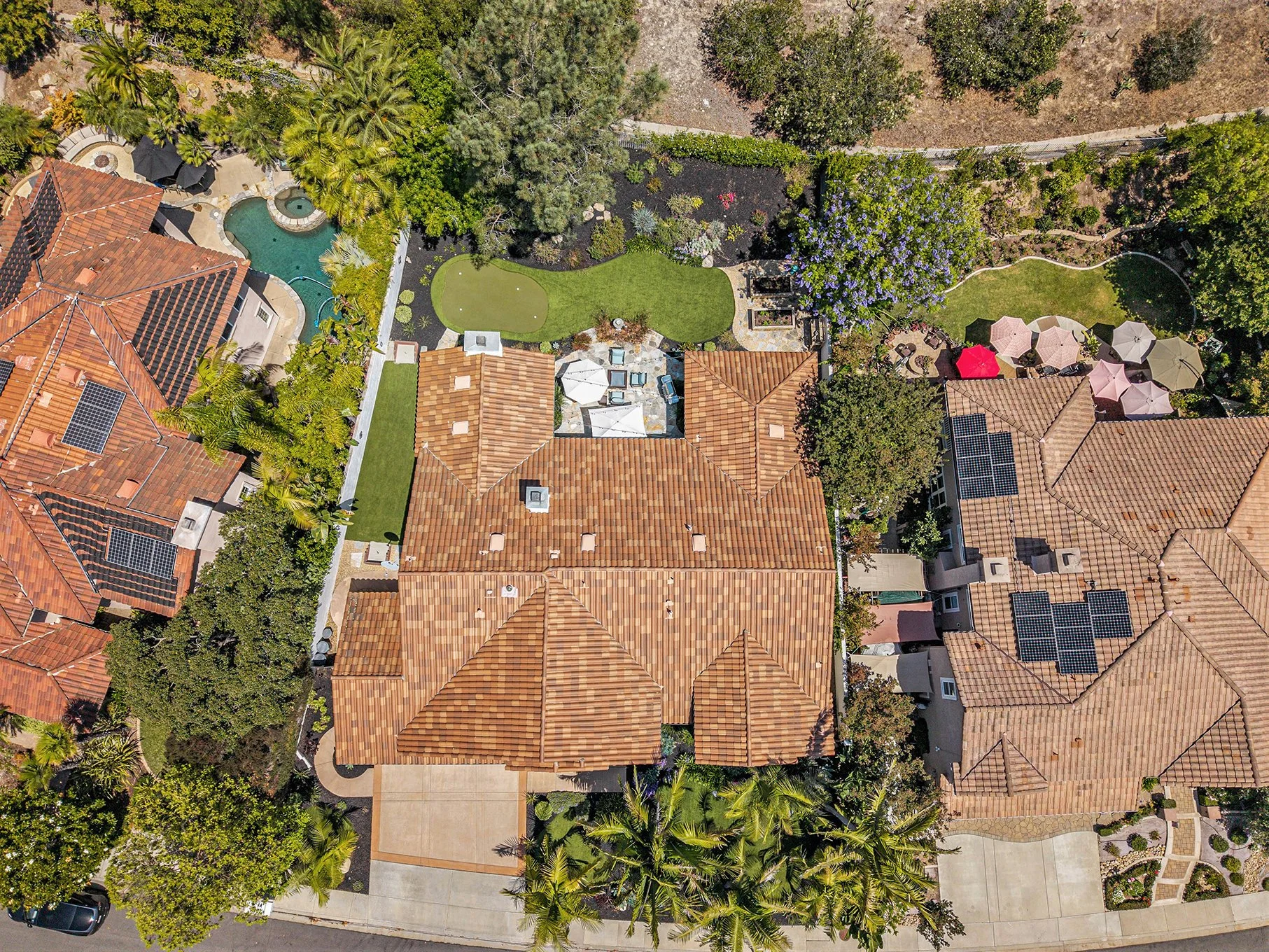 Aerial view of a residential backyard with a swimming pool, multiple patio areas, and landscaped gardens surrounded by neighboring houses with tiled roofs and solar panels.