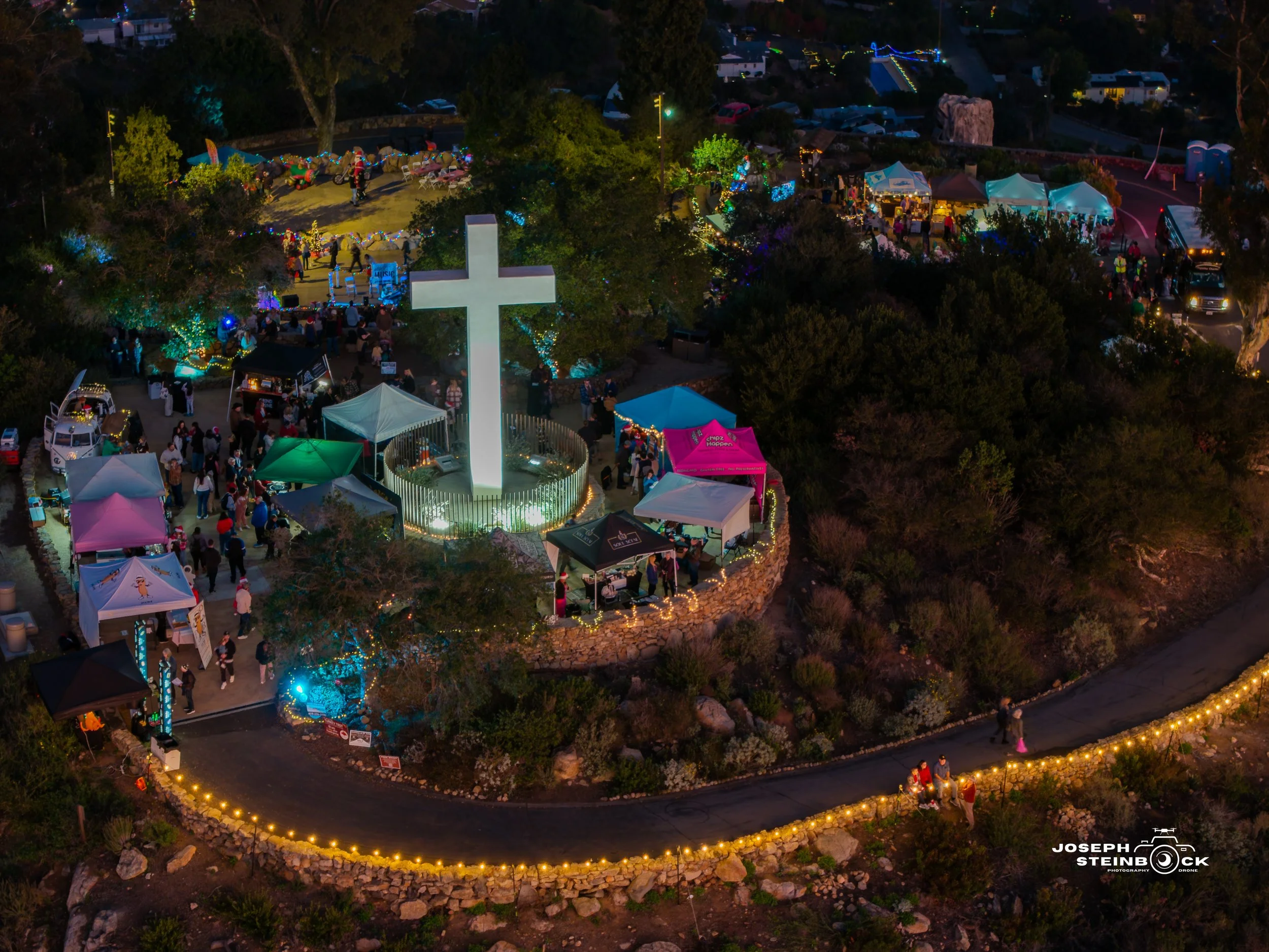 A night scene at a community gathering in a park with many tents, booths, and string lights. A large illuminated cross is in the center surrounded by people. Tents and booths are arranged around the cross, with some people walking along a winding, li