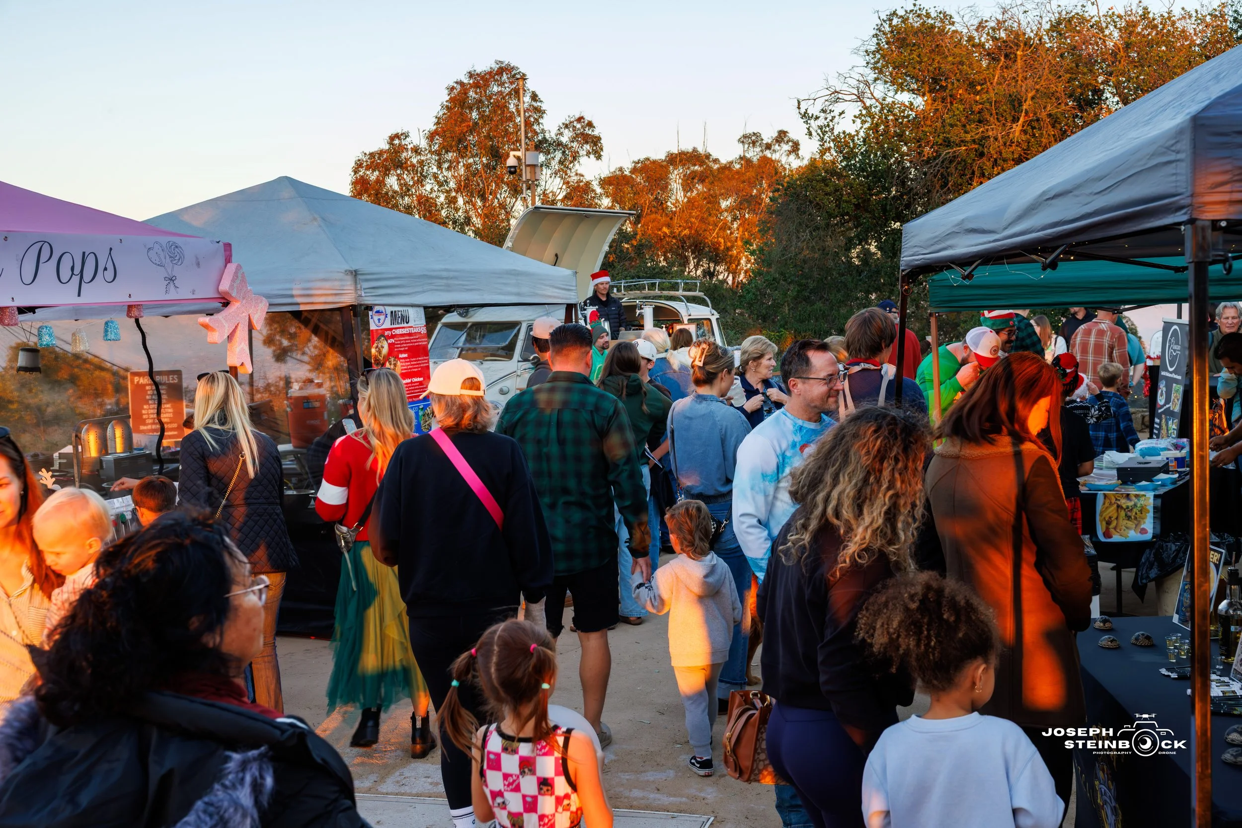 Crowd of people at an outdoor market or festival with food stalls, decorated for Christmas, during sunset, with trees in the background.