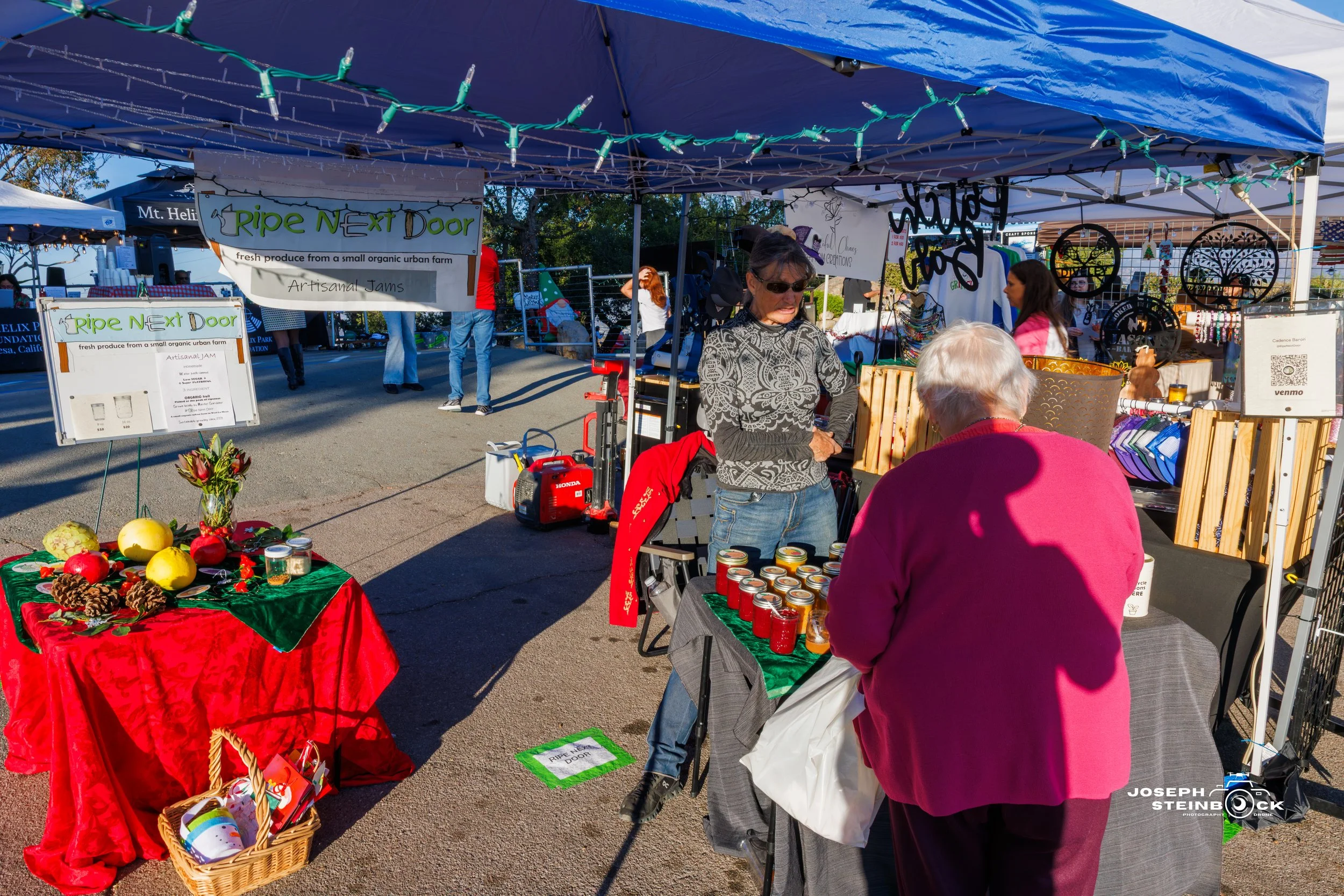 At an outdoor market stall, a woman in sunglasses sells jars of jam to an elderly customer with white hair in a pink sweater. The stall has a sign reading 'Ripe Next Door' and several produce and crafts on display, including a decorated table with pi