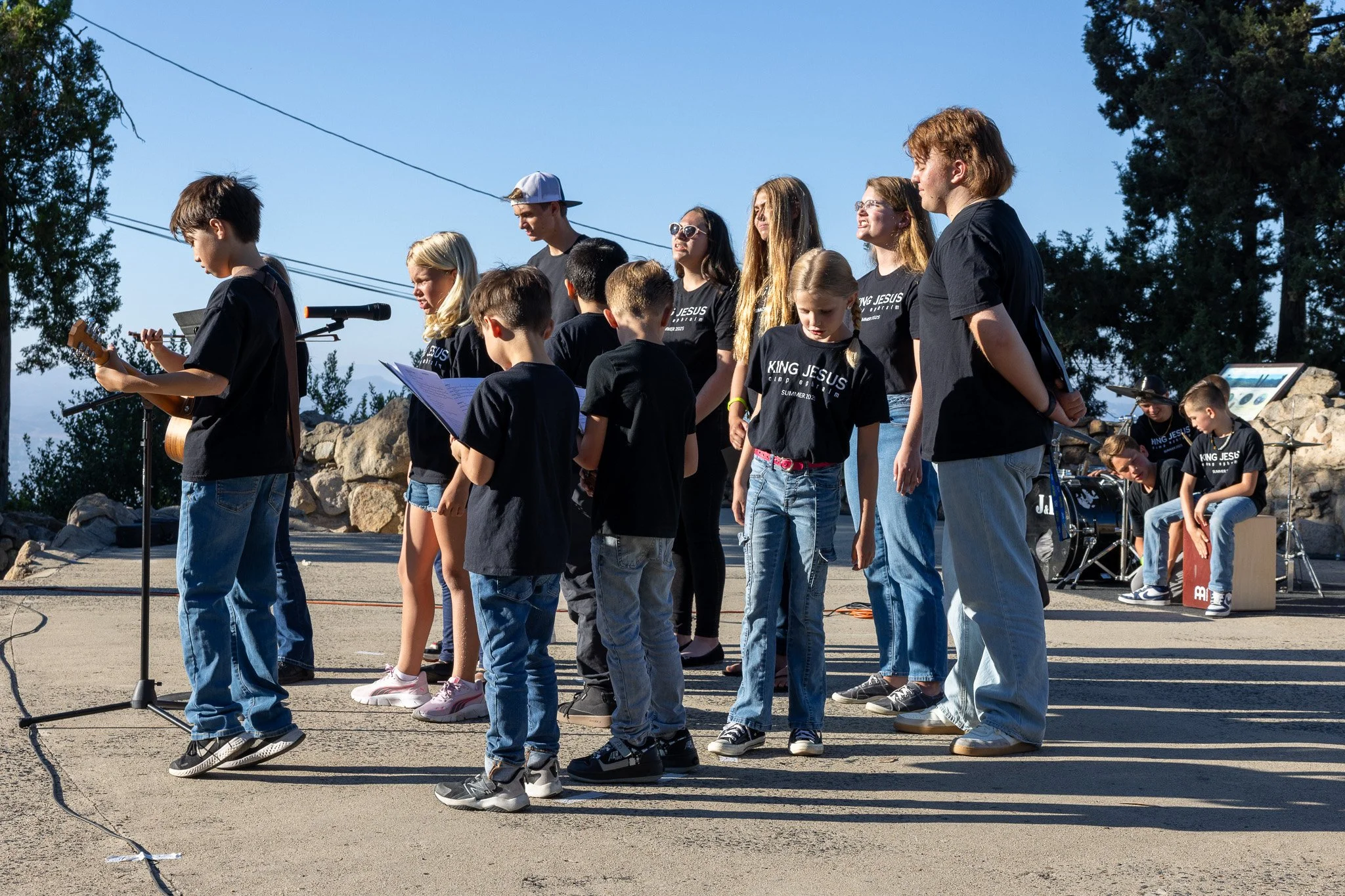 A group of children and teenagers wearing black t-shirts with 'King Jesus' written on them, standing outdoors on a stage with rocks and trees in the background, participating in a musical performance with some holding a music sheet and others playing