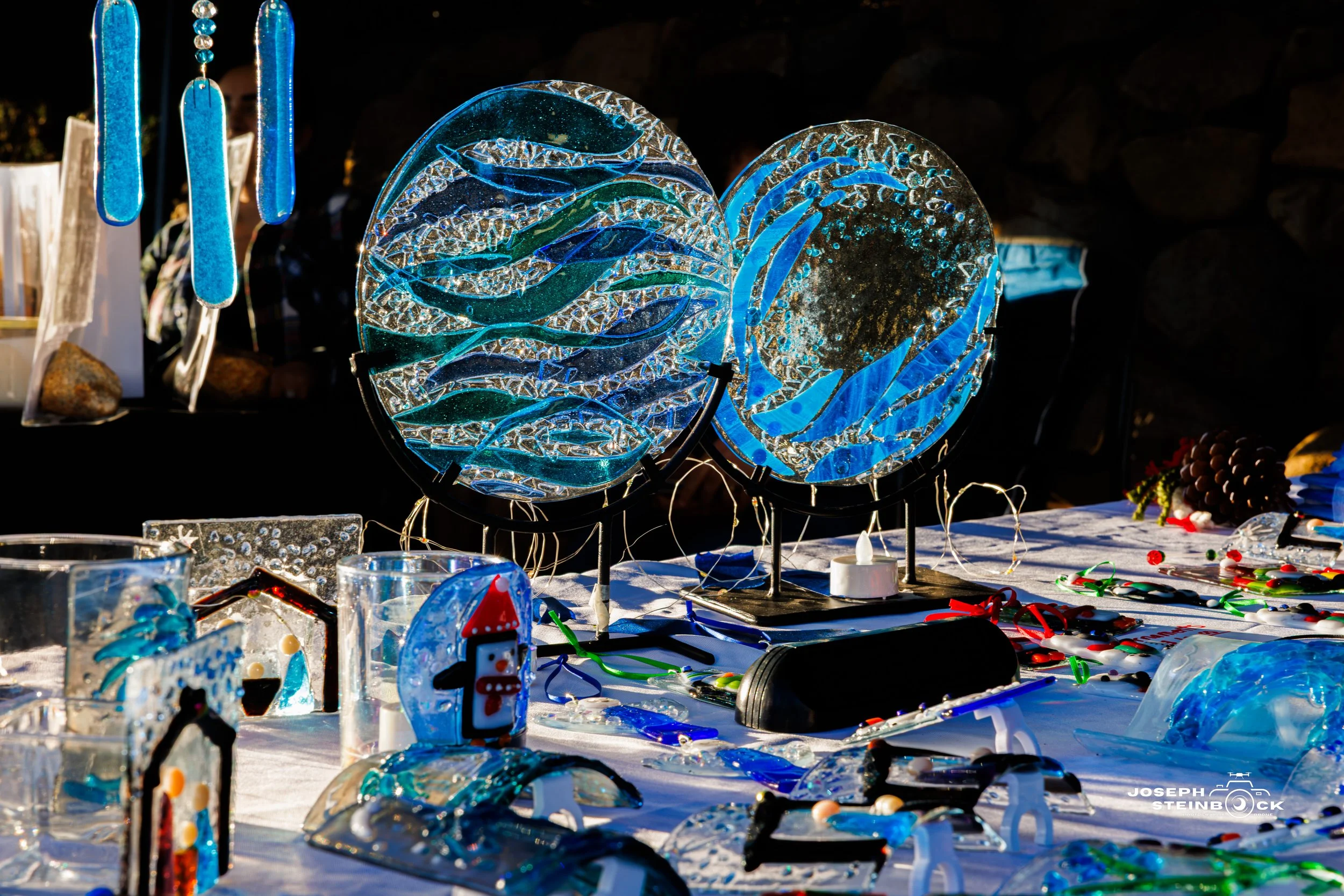 Glass art display featuring two circular glass plaques with blue and clear abstract patterns, illuminated on black stands. The table has various glass crafts including small sculptures, ornaments, and holiday-themed items, with other colorful glass p